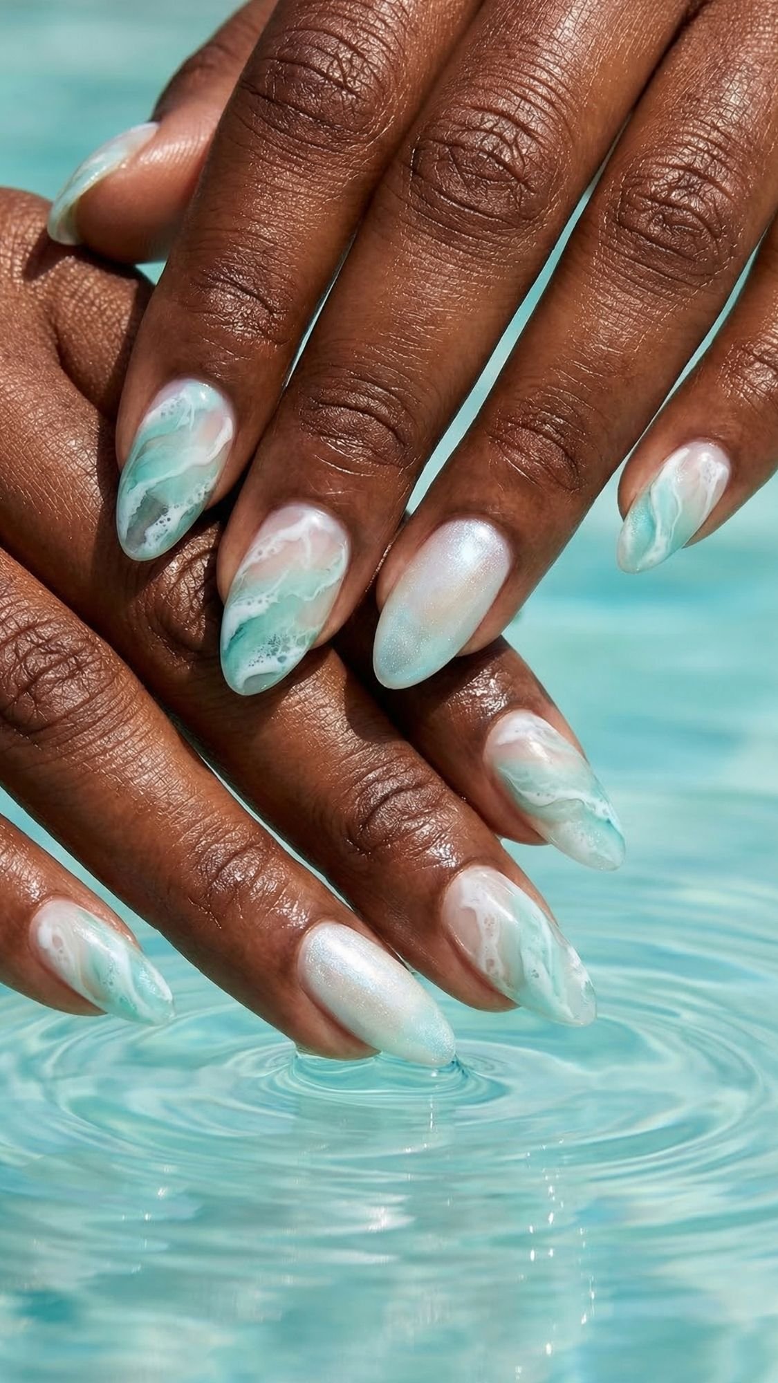 Two hands with almond-shaped nails feature aqua and white marbled nail art, resembling ocean waves—perfect beach nails. The fingertips gently touch the surface of clear blue water, creating small ripples.