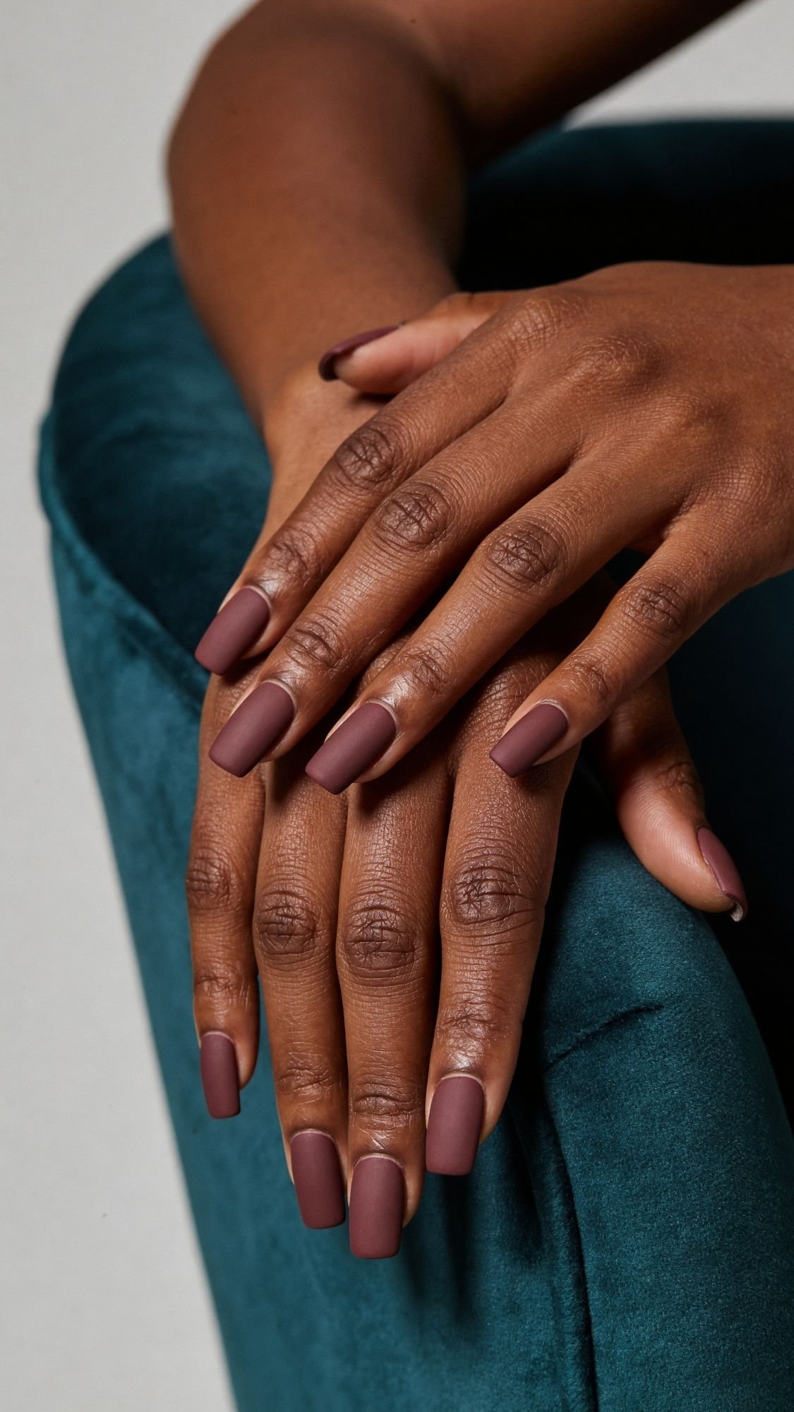 Two hands with dark brown matte, square-shaped chic nails rest gently on a teal velvet surface. The skin tone is deep brown, and the professional nail design appears well-groomed, stylish, and work friendly.