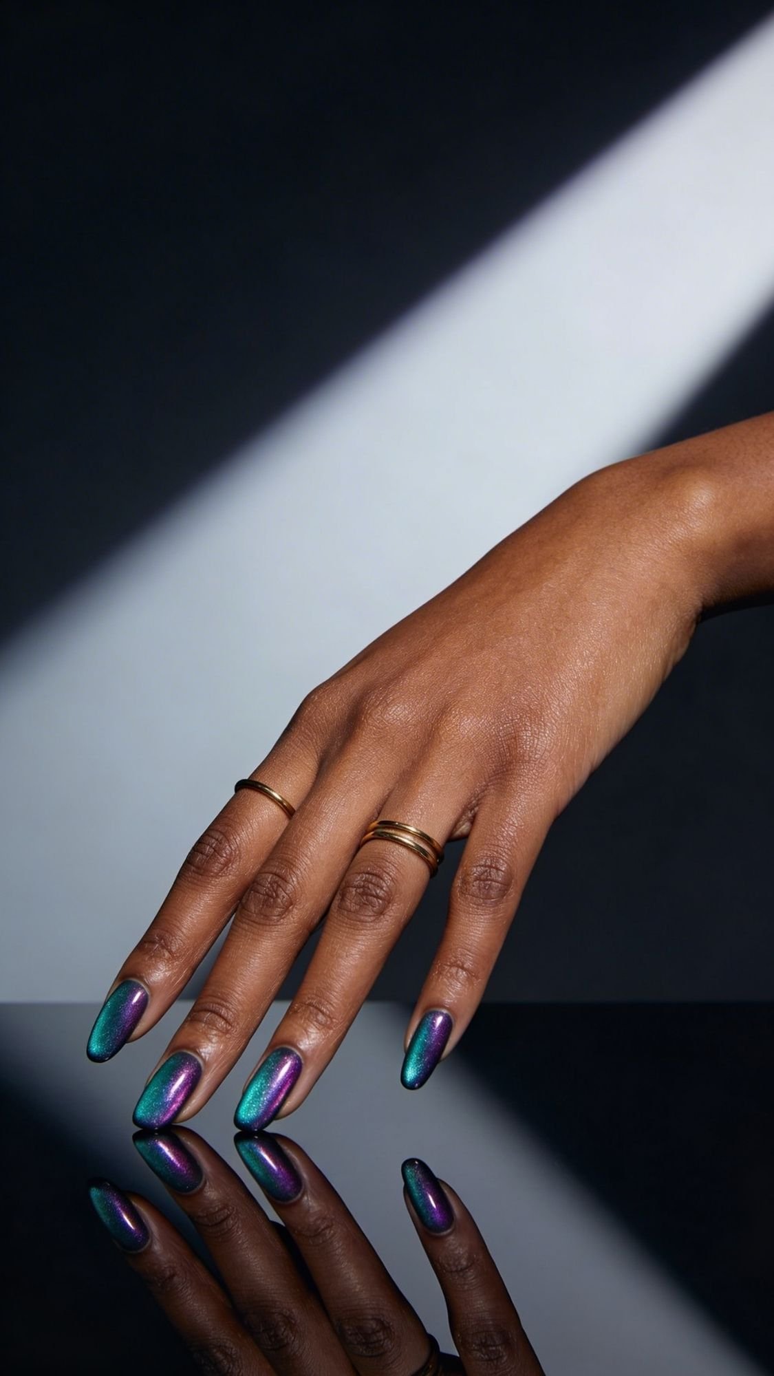 A hand with metallic, multicolored chrome nails rests on a reflective black surface. The person wears two thin gold rings, channeling serious 16 Chrome Nail Ideas vibes as dramatic lighting casts diagonal shadows in the background.