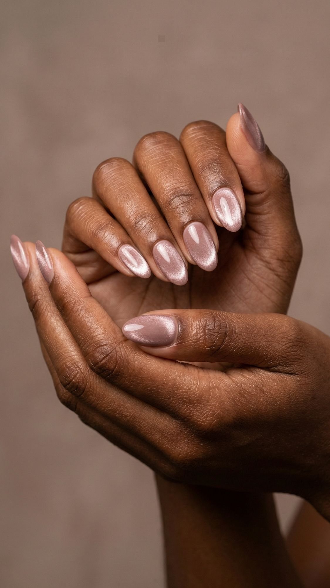 A close-up of two hands with dark skin, featuring neatly manicured almond nails painted with a shiny, pale pink polish. The flattering nails are gracefully posed against a neutral background.