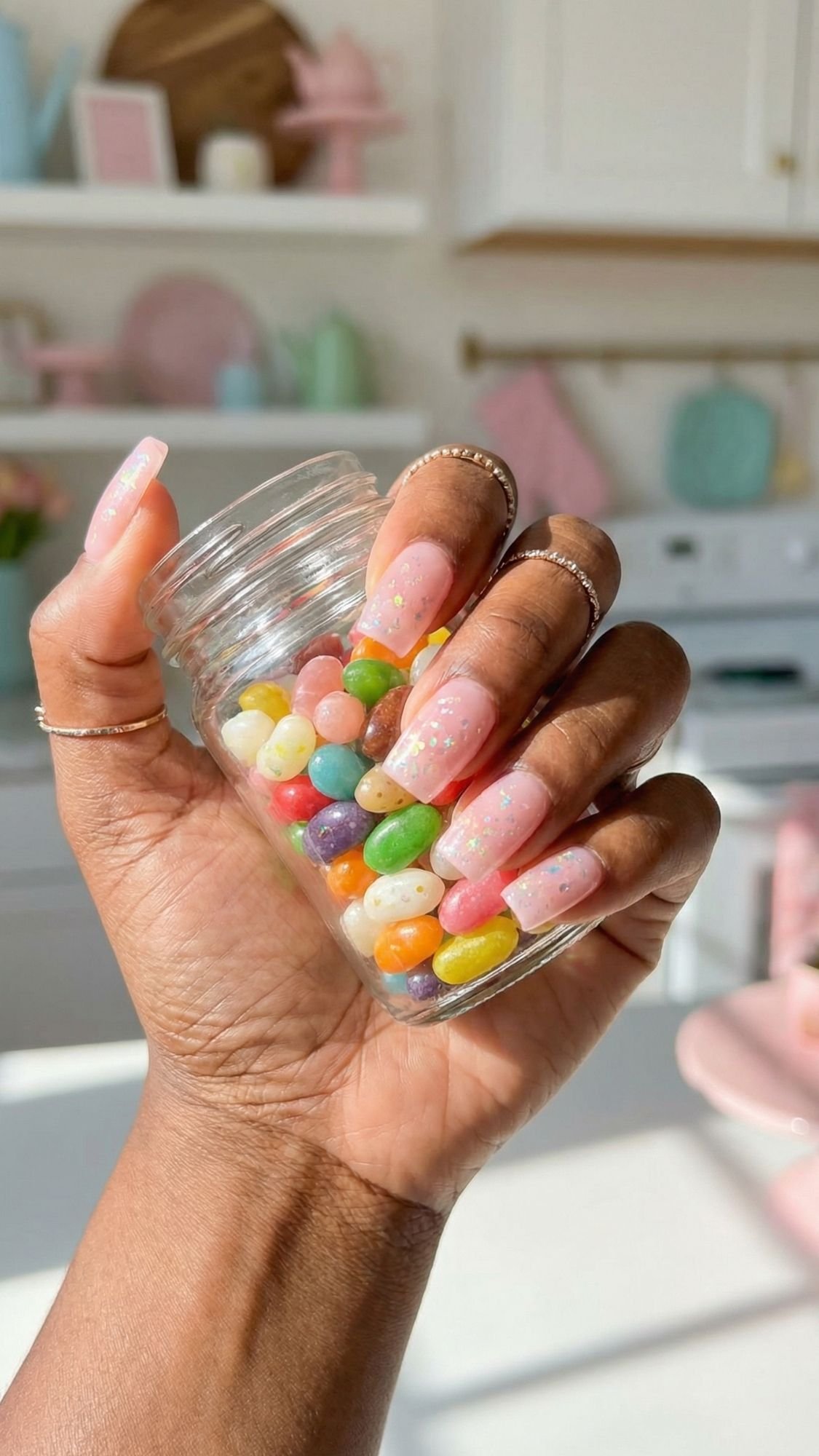 A hand with long pink sparkle nails holds a small glass jar filled with colorful jelly beans in a bright, pastel-colored kitchen.