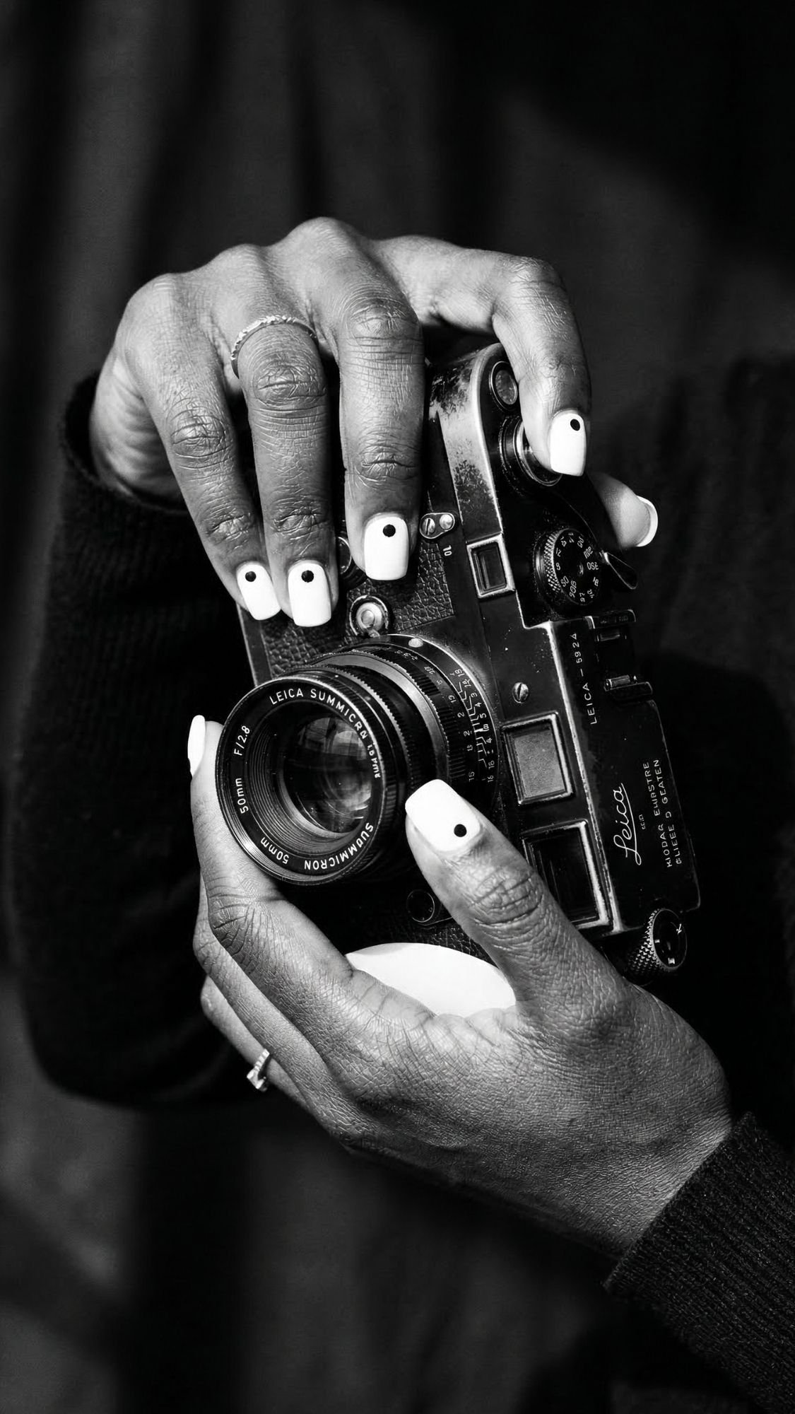 A close-up black-and-white photo shows two hands with elegant nails holding a vintage Leica camera. The person’s short nails are painted white with small black dots, and they are wearing a thin ring on one finger.