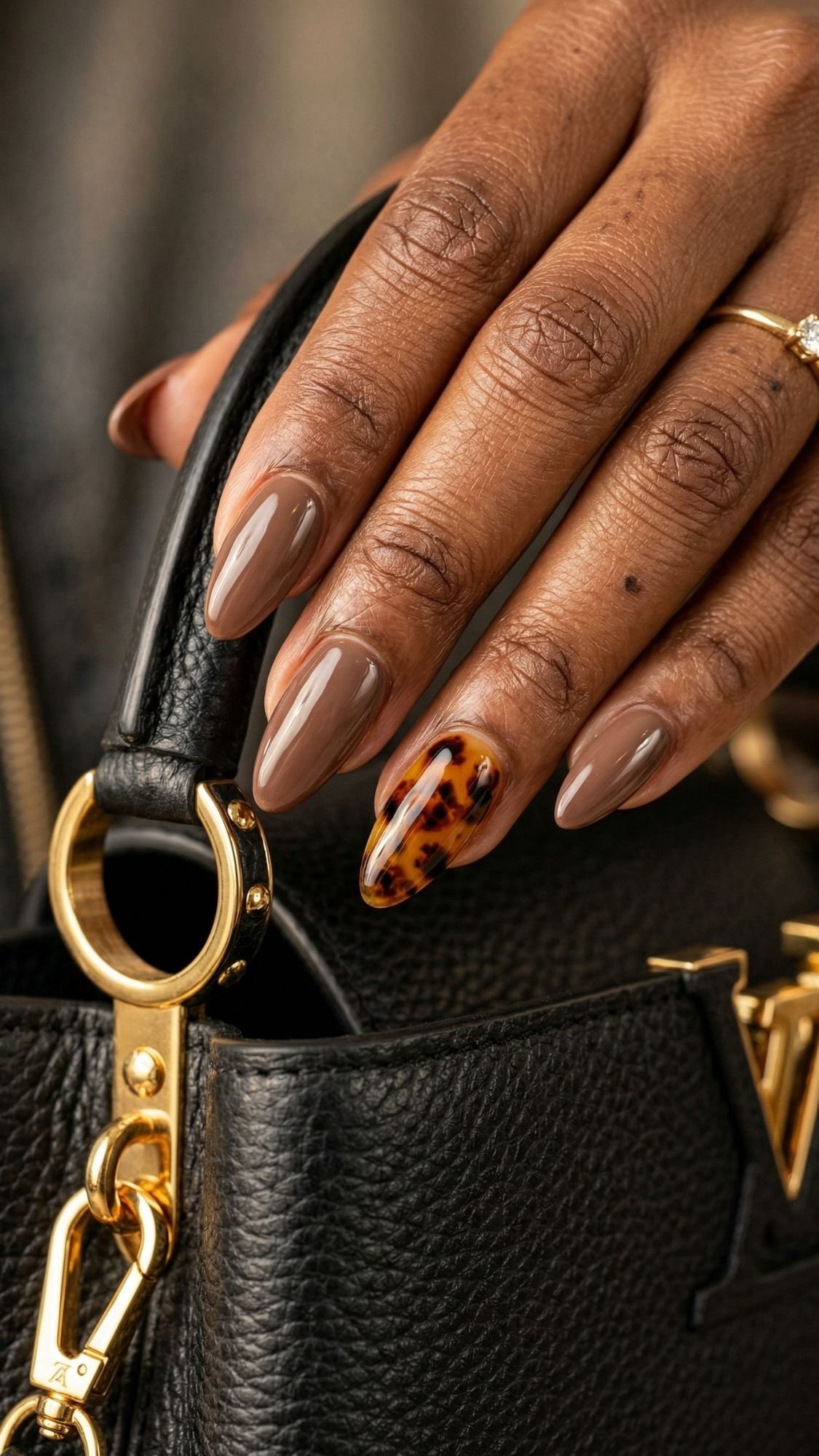 A hand with almond-shaped chic nails painted in glossy taupe, featuring one accent tortoiseshell nail, holds a black textured leather bag with gold hardware—perfect inspiration for professional nail designs and work friendly nails.