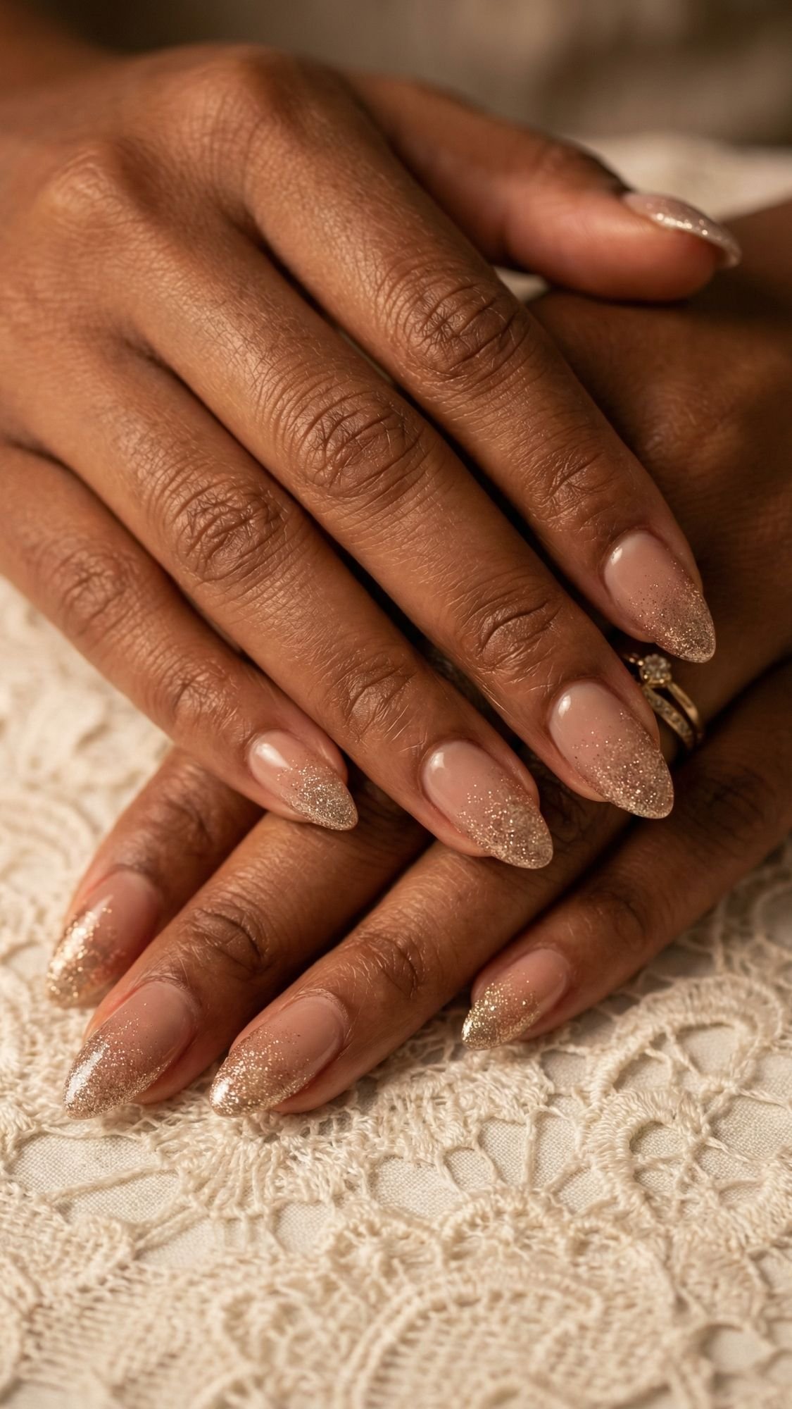 Two hands with long, almond-shaped nails featuring a nude base and glittery gold tips, resting on white lace. The person wears gold rings on one finger—perfect Sparkle Nails inspiration for your next Holiday Nail Ideas.