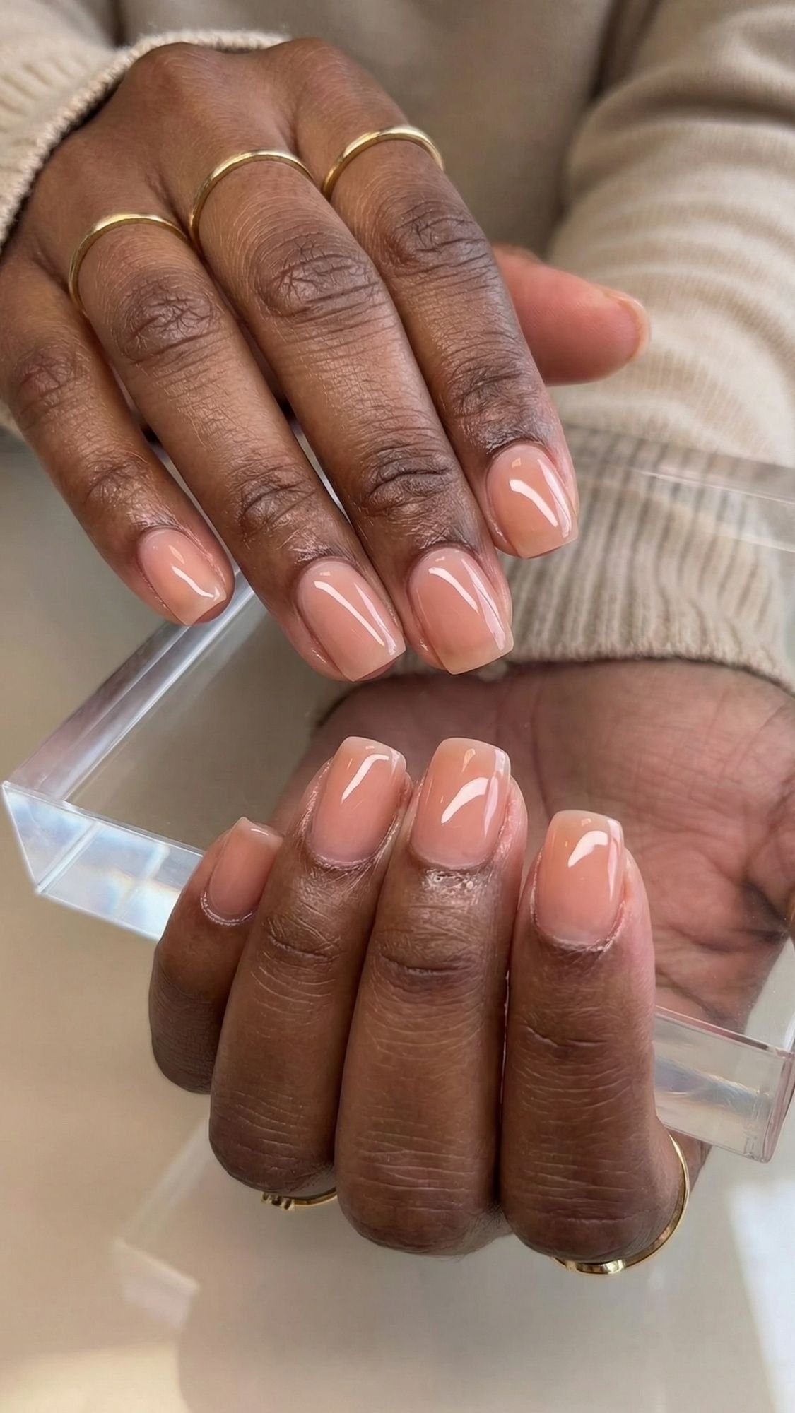 Close-up of hands with medium-length, square-shaped nails painted in a glossy nude color—perfect wedding guest nails. The person wears gold rings and a beige sweater, holding a clear acrylic block.