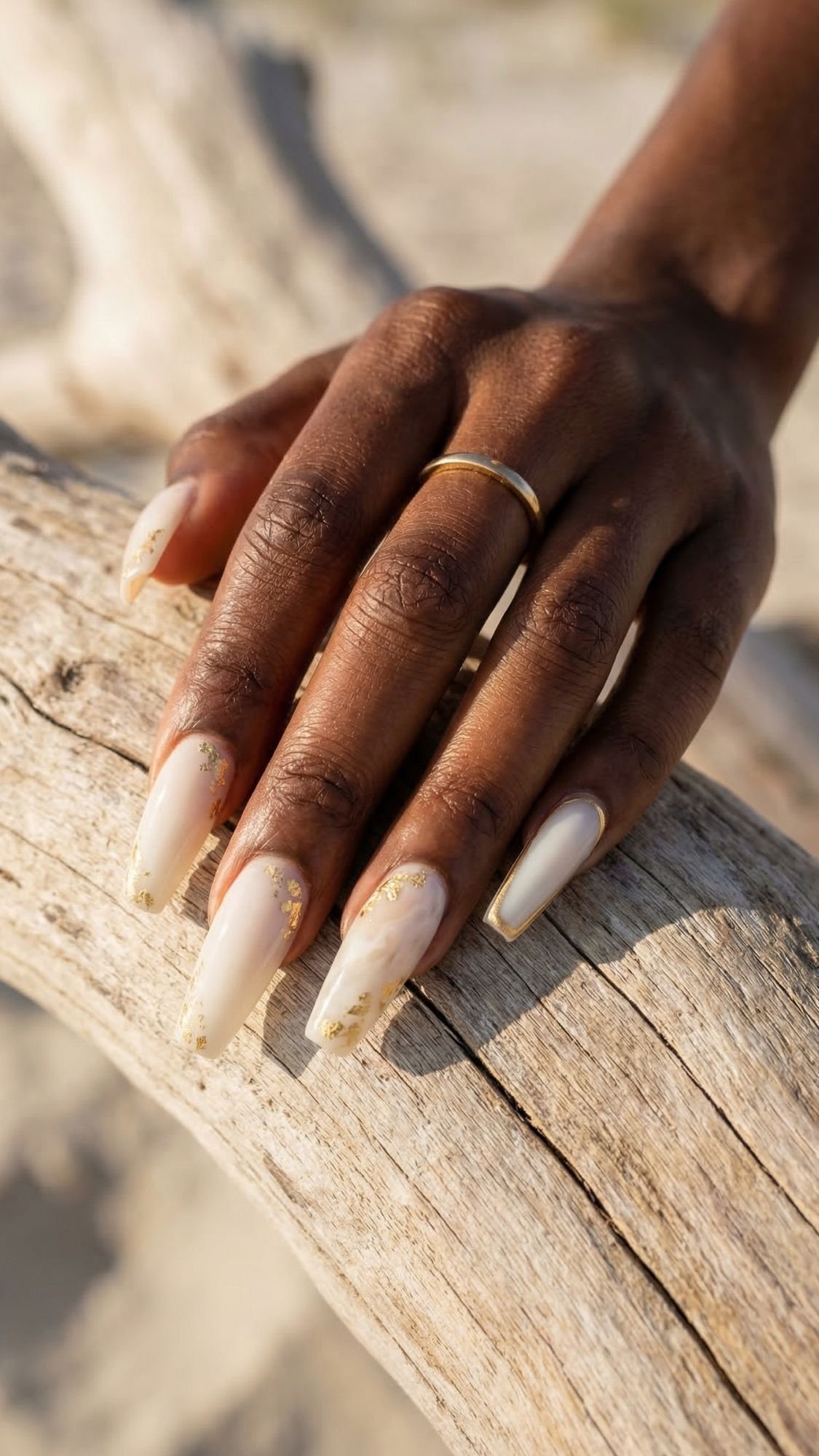 A hand with long, almond-shaped nails featuring a soft nude polish with gold accents—perfect for beach nails—rests on driftwood. The person is wearing a simple gold ring on their finger.
