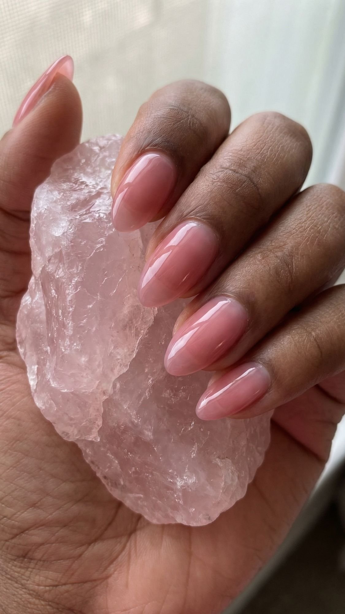 A hand with long, glossy, pale pink almond-shaped nails holds a large, rough rose quartz crystal against a softly lit background—perfect clean girl nails for an effortlessly polished look.