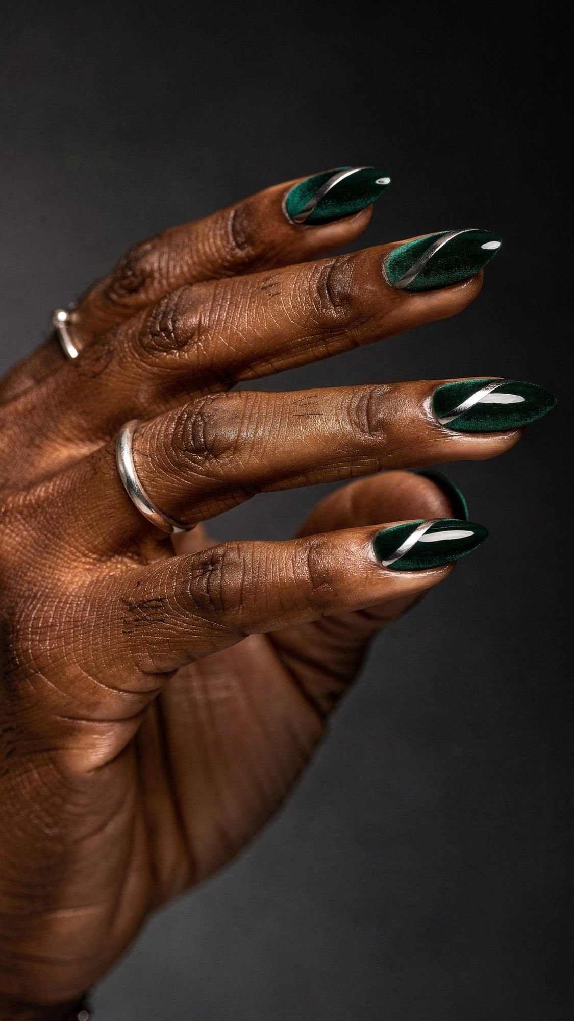 A hand with dark skin and silver rings displays manicured, shiny nails painted dark green with a glossy finish and a white diagonal stripe design on each nail, against a dark background—a subtle take on selfie nails.