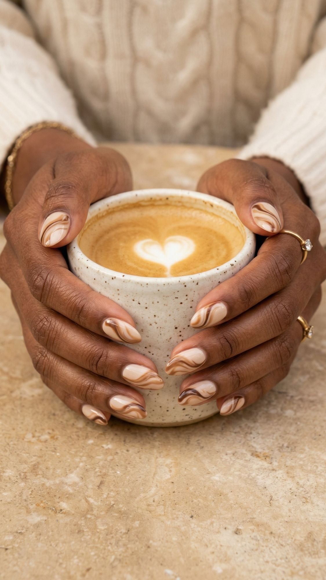 A person with brown skin and elegant, almond nails featuring marble-patterned nail designs holds a speckled ceramic cup of latte with heart-shaped foam art, resting on a beige stone surface.