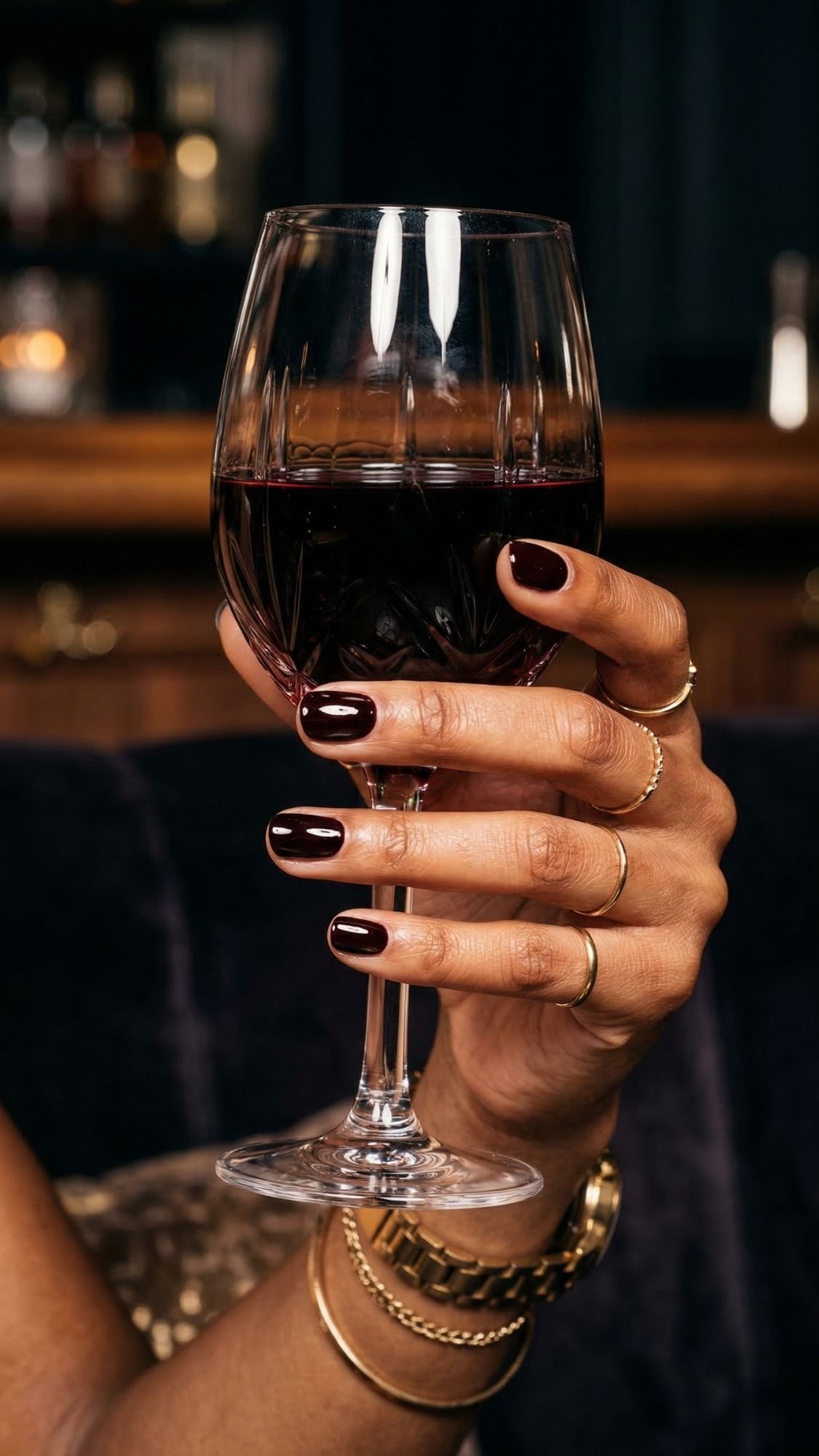 A hand with elegant short nails, polished dark and adorned with gold rings, holds a glass of red wine against a blurred bar setting.