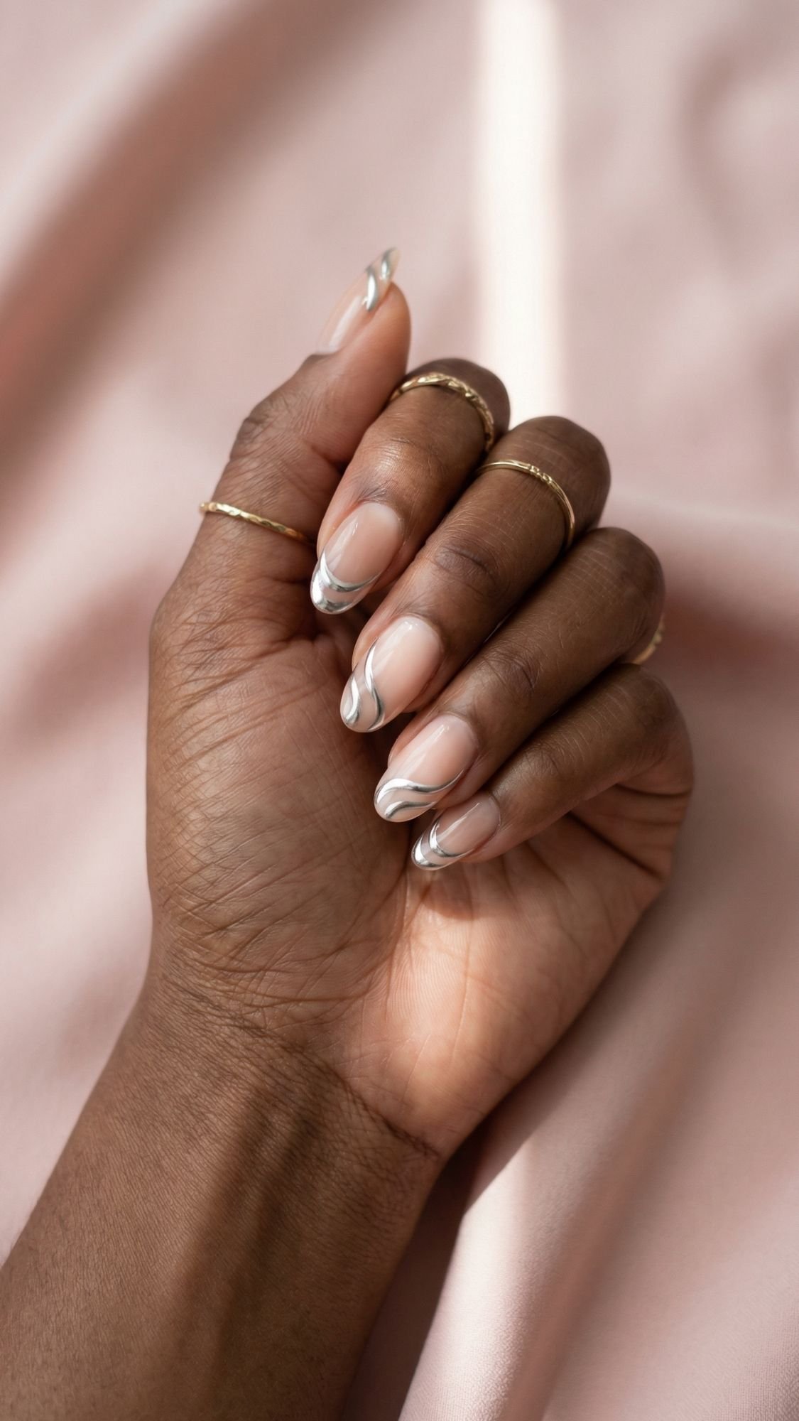 A hand with gold rings displays fingernails painted in a nude polish with stylish white and silver chrome nails abstract tips, resting on a soft, light pink fabric background.