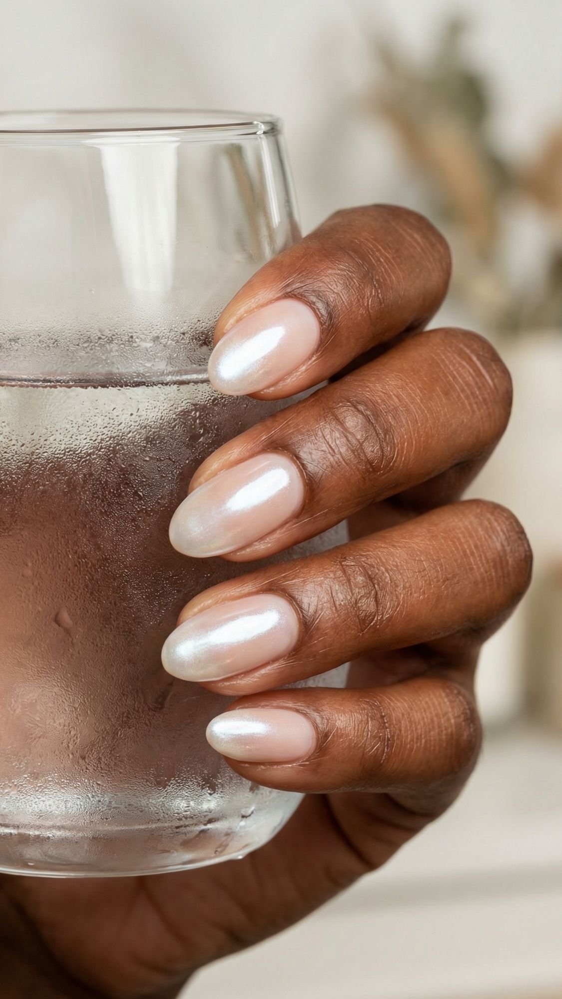 A hand with chic, pearly, glossy, almond-shaped nails holds a clear glass of water with condensation. The skin tone is medium to dark brown. The softly blurred background adds to the stylish, work friendly nails aesthetic.