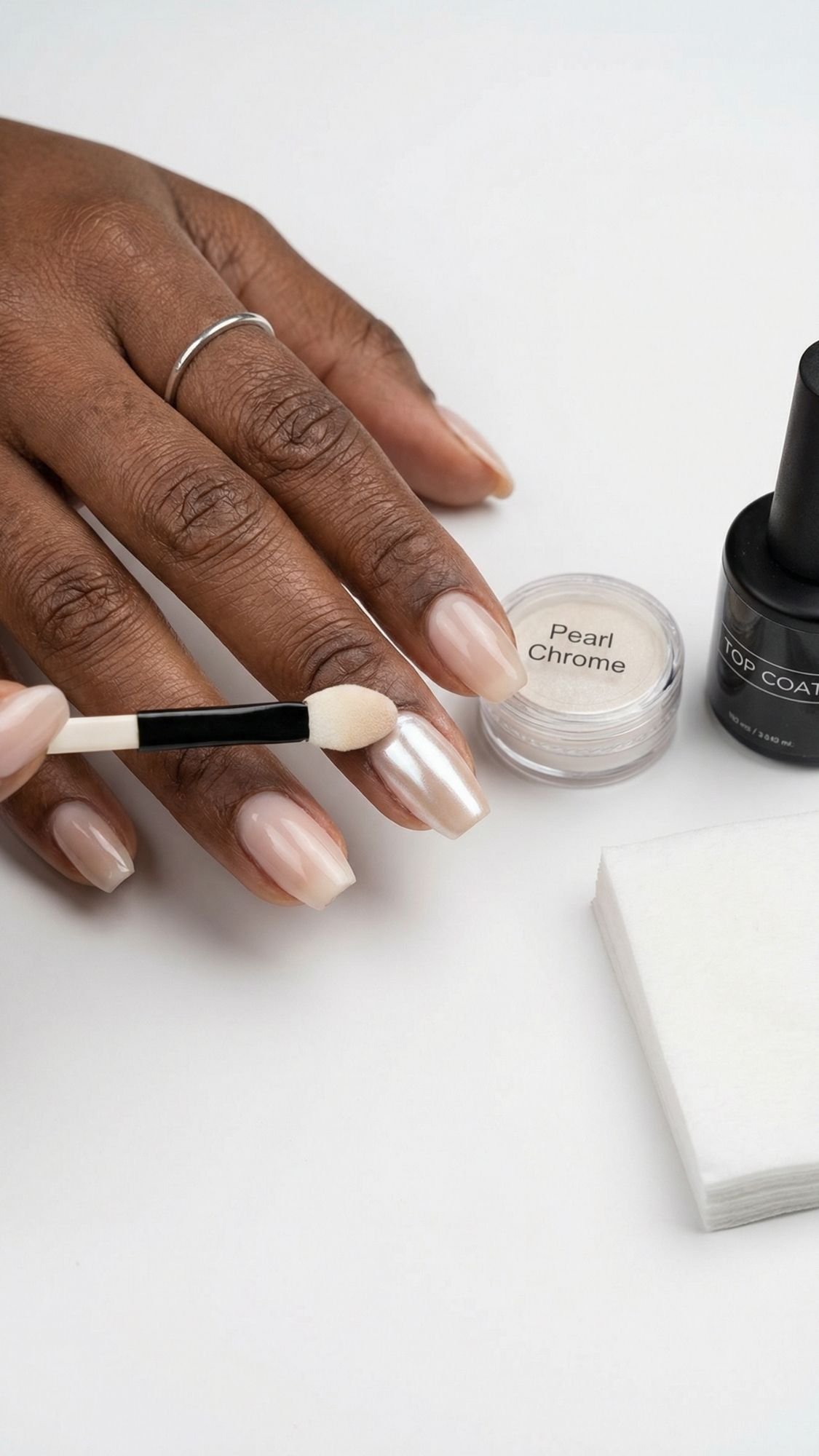 A hand with neatly manicured nails is having pearl chrome powder applied for stunning chrome nails. Nearby are a jar labeled Pearl Chrome, a bottle of top coat, and a stack of white nail wipes—perfect for light catching nails in every selfie.