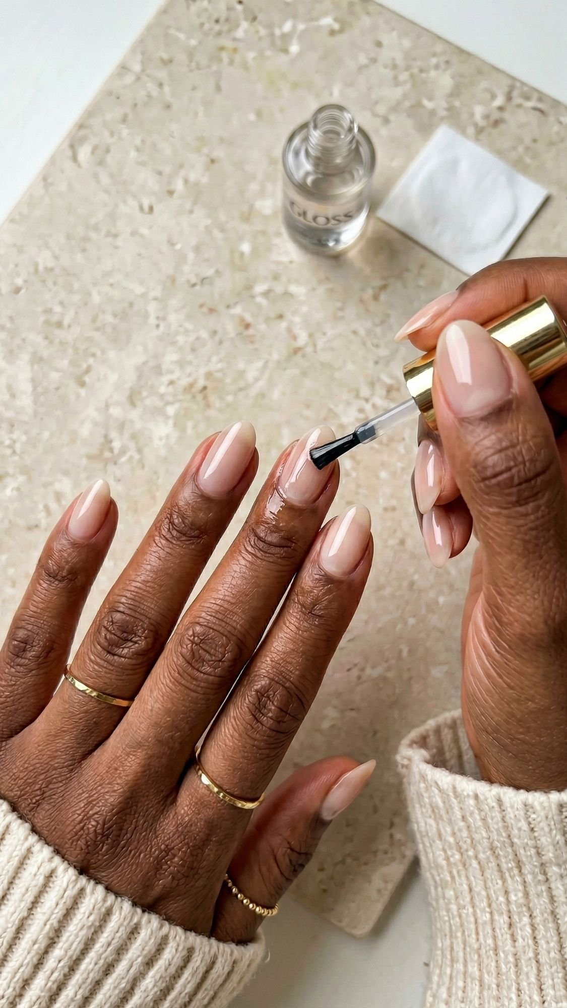 A person with medium brown skin and neatly manicured almond nails is applying clear polish. They wear several gold rings and a cream sweater. A nail polish bottle and cotton pad sit on a beige surface, highlighting flattering nail shapes.