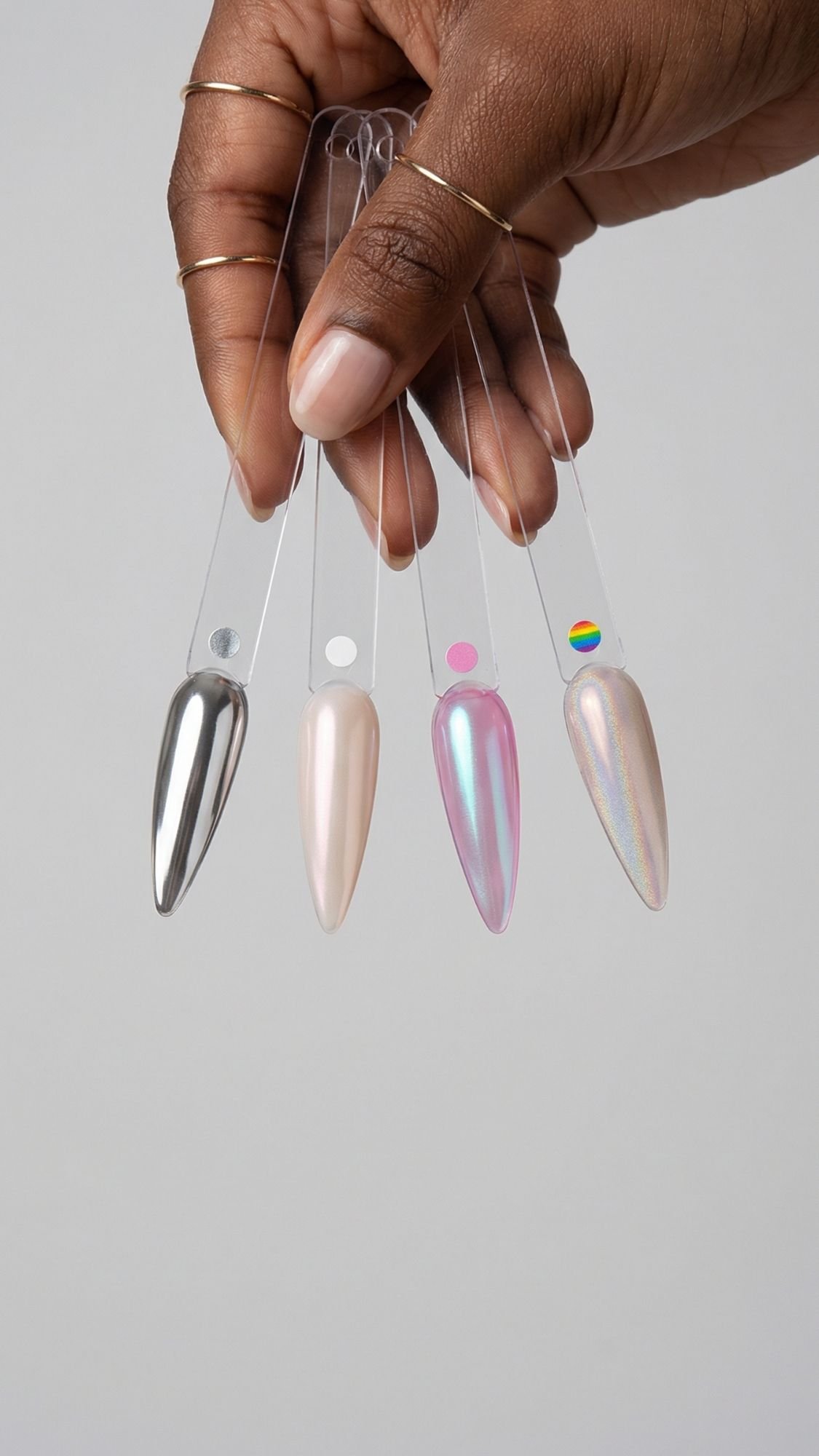 A hand with gold rings shows off four nail polish swatches in metallic and iridescent shades, including silver, pearl, pink, and selfie-ready chrome nails, all set against a plain light gray background.