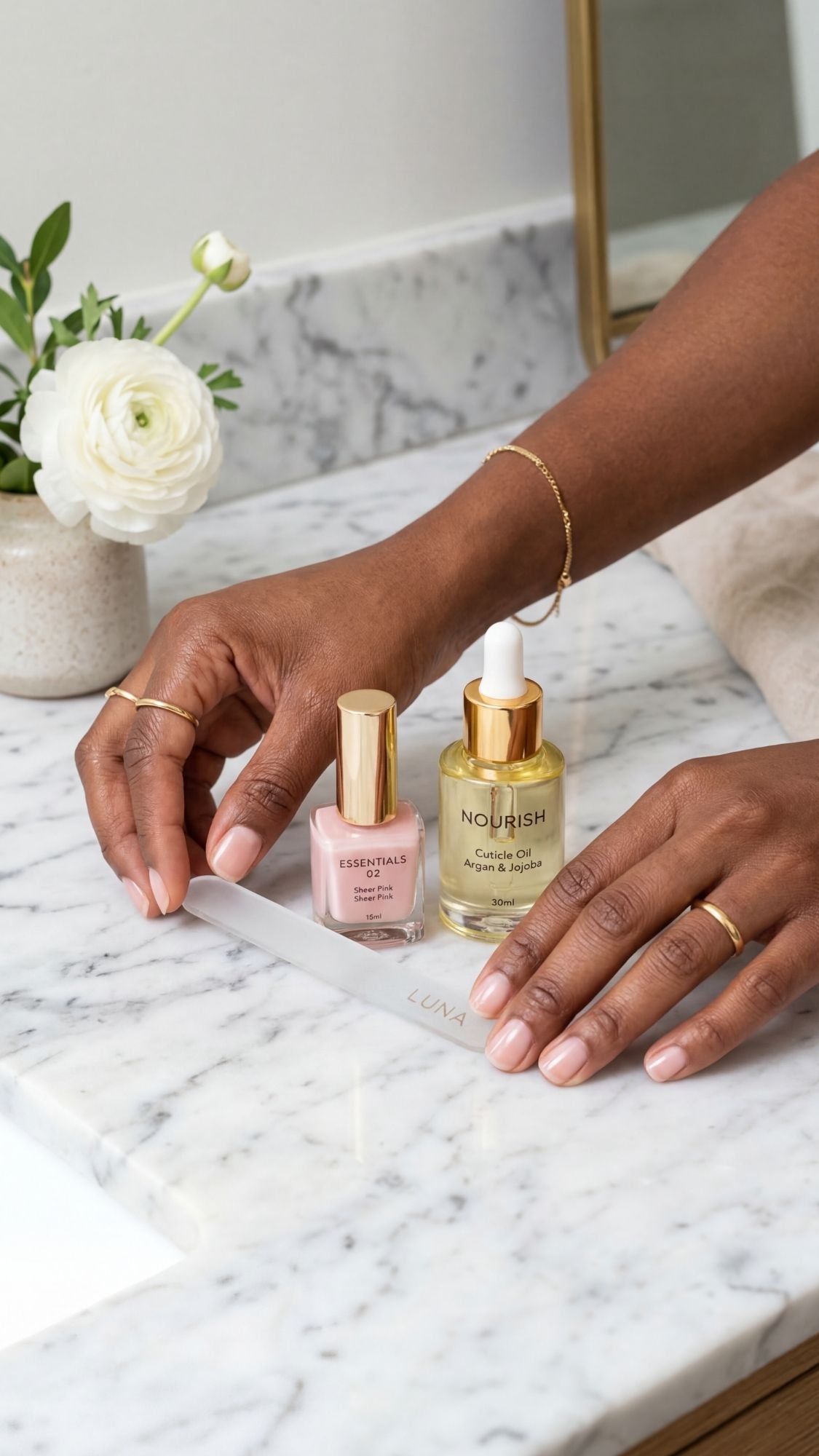 A person’s hands place a white strip on a marble countertop next to a pink nail polish bottle—perfect for elegant nails or short nails—with a gold-capped facial oil, white flower, and towel in the background.