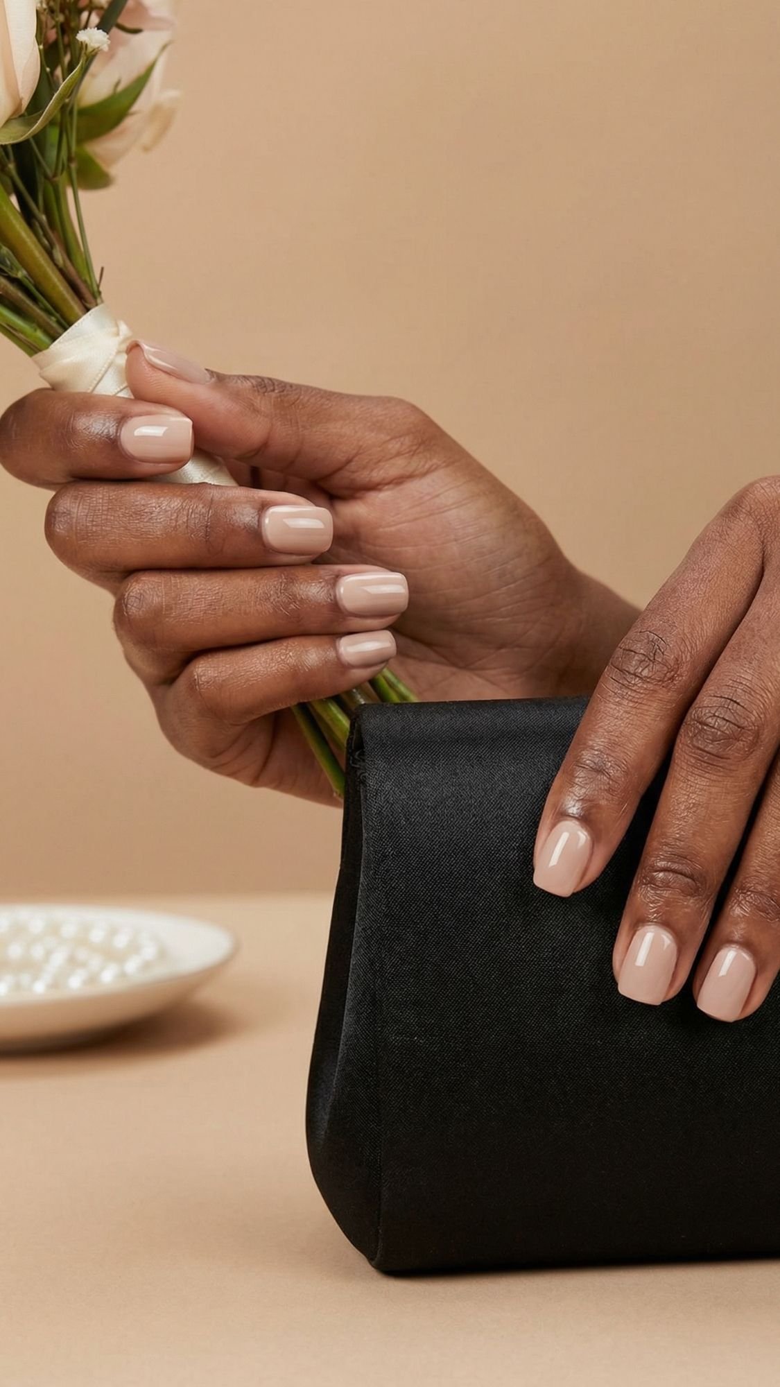 A close-up of hands with neatly manicured nude nails, perfect wedding guest nails, one hand holding a small bouquet of flowers and the other resting on a black clutch purse, against a beige background.