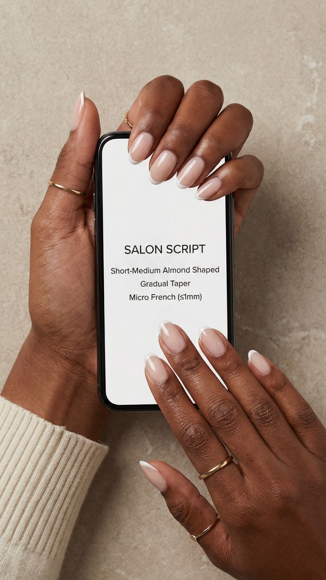 A person with almond-shaped, micro French manicured nails holds a phone displaying text about flattering nail designs on a neutral surface. The person wears gold rings and a cream sweater.