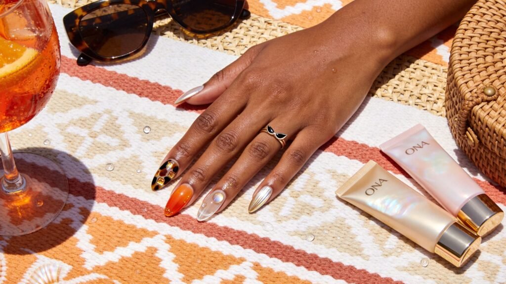 A hand with colorful, patterned brunch nails rests on a patterned towel next to sunglasses, a woven bag, a drink with orange slices, and two ONA skincare tubes. The nails feature orange, gold, and tortoiseshell designs.