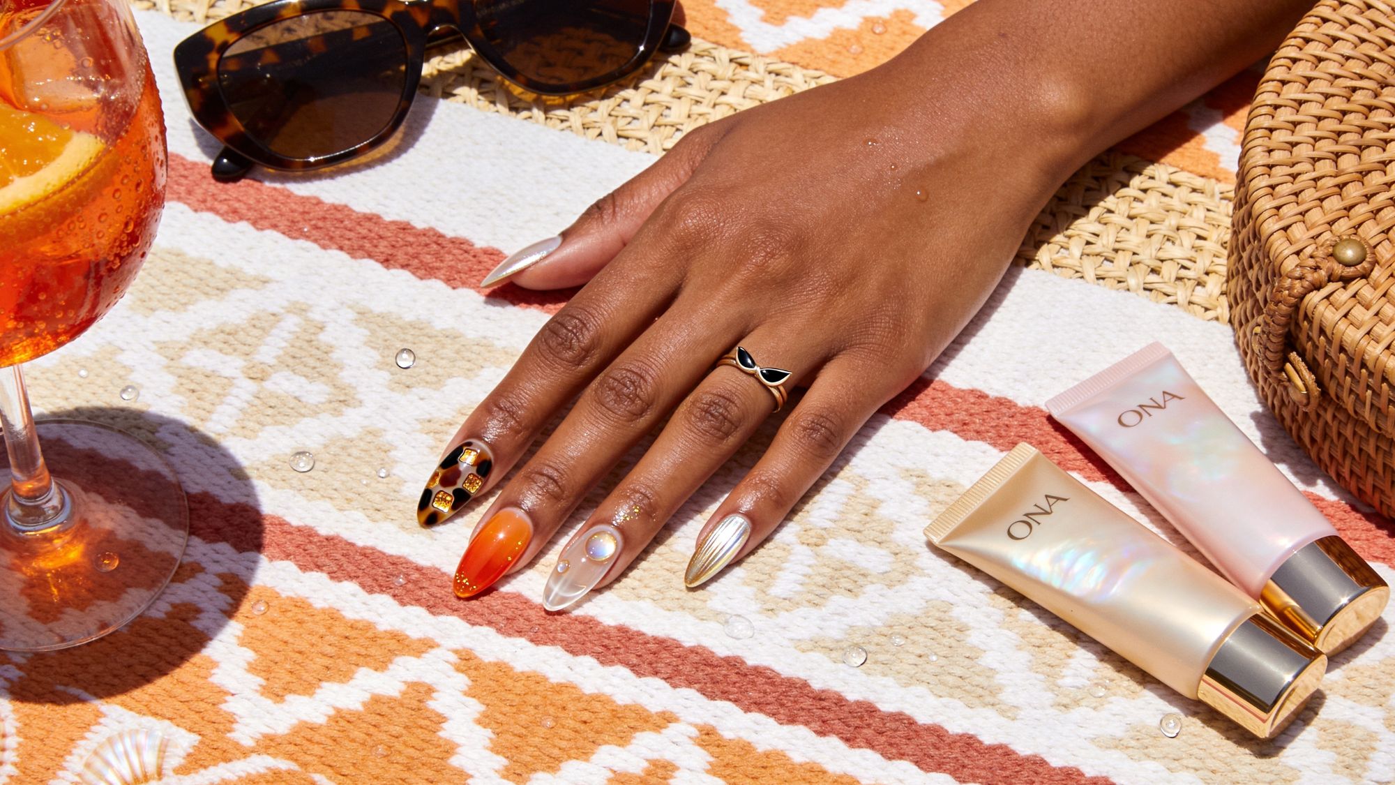 A hand with colorful, patterned brunch nails rests on a patterned towel next to sunglasses, a woven bag, a drink with orange slices, and two ONA skincare tubes. The nails feature orange, gold, and tortoiseshell designs.