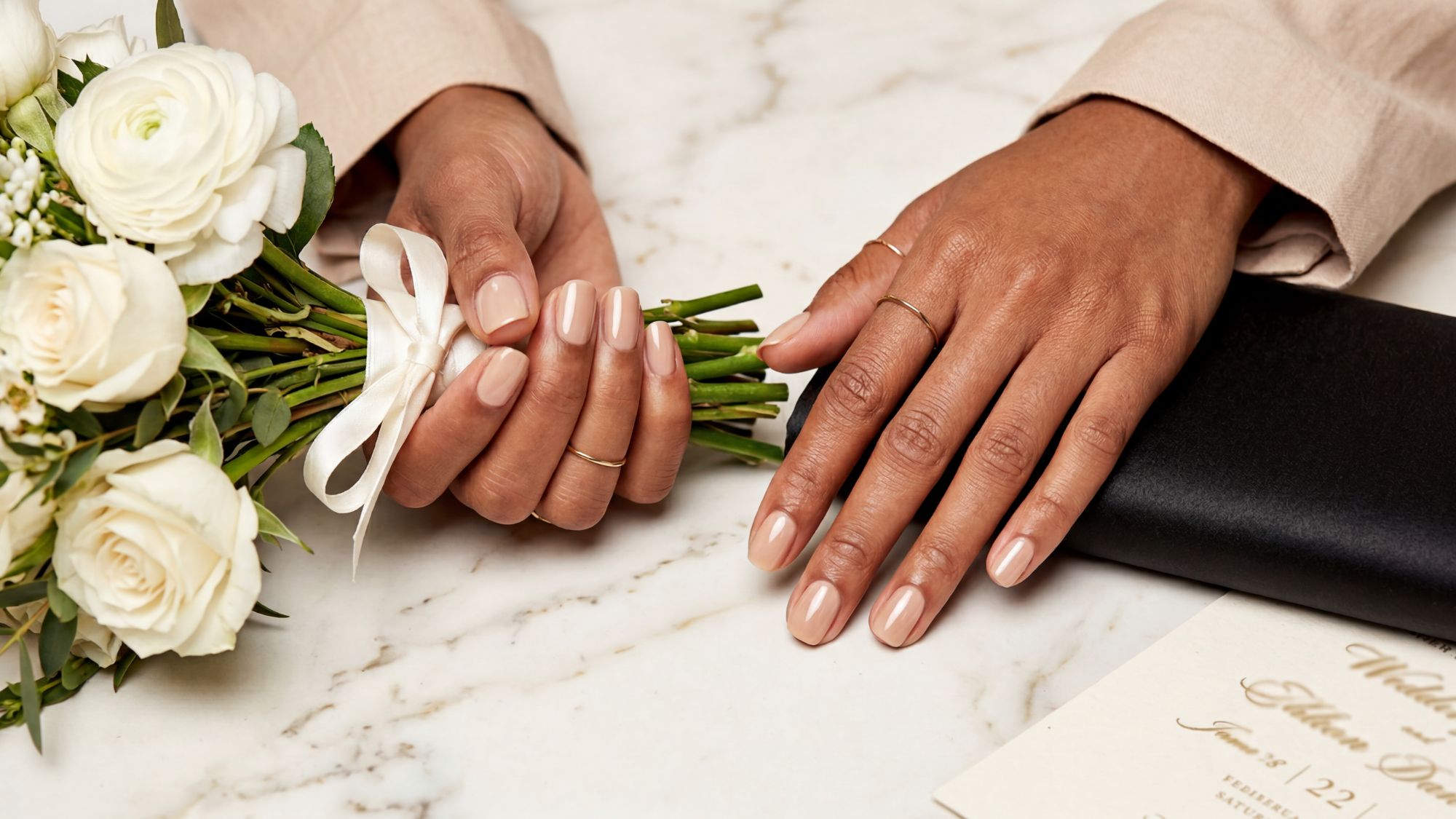 Close-up of hands with nude wedding guest nails holding a small bouquet of white roses and greenery. One hand rests on a marble surface near wedding invitations and a black envelope, perfect nails for any dress.