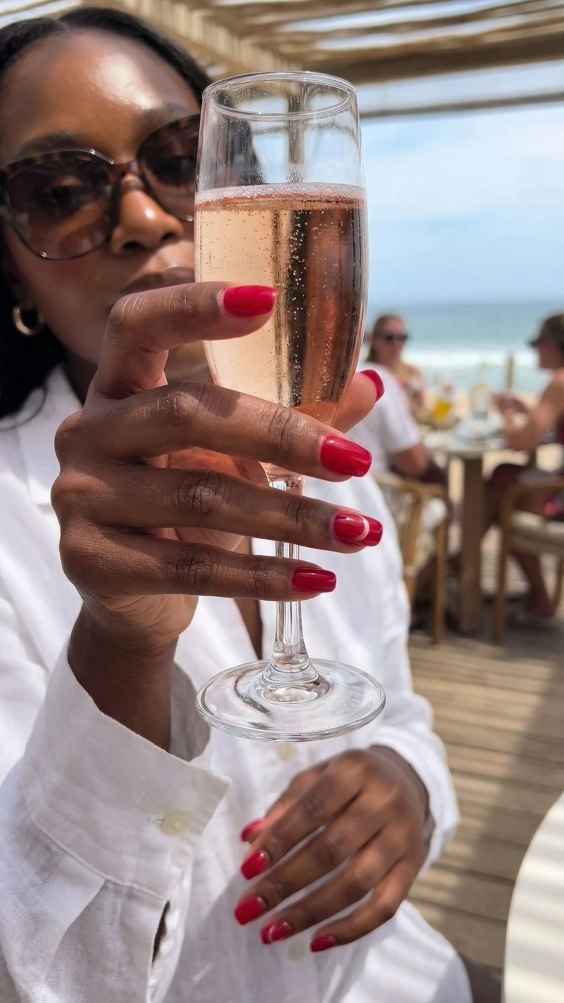 A woman wearing sunglasses and a white shirt holds up a glass of sparkling rosé, showing off her chic red brunch nails. She is seated at an outdoor restaurant by the beach, with people and the ocean in the background.