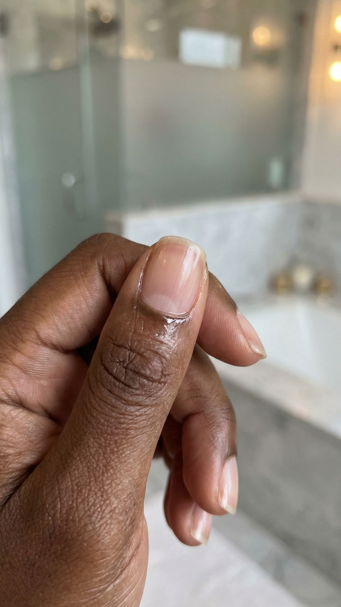 A close-up of a person’s hand with a peeling cuticle and dry skin, in front of a blurred bathroom background with a bathtub and frosted glass shower—highlighting the contrast to the Everyday Look of polished nails.