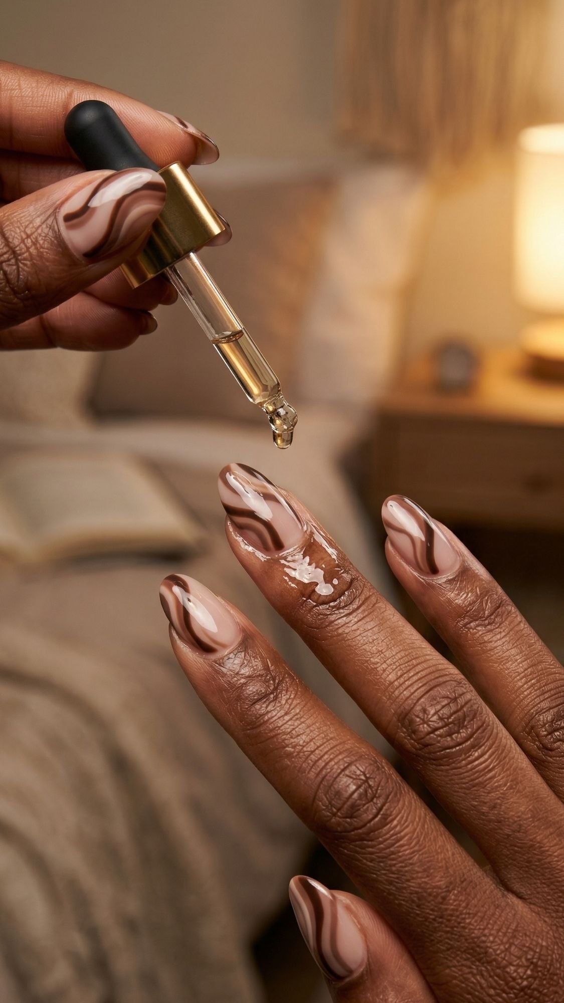 A hand with brown and white marbled, work friendly nails has cuticle oil applied by dropper. The softly lit background reveals a bed, a lamp, and a nightstand topped with an open book.