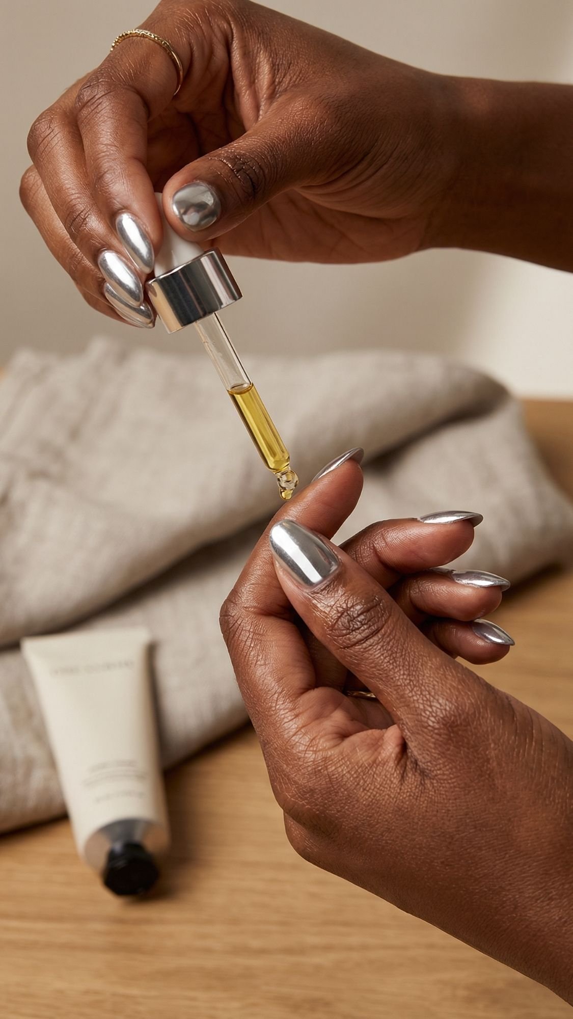 A person with chrome nails uses a dropper to apply yellow facial oil onto their hand. A beige fabric and a tube of skincare product are on a wooden surface in the background.