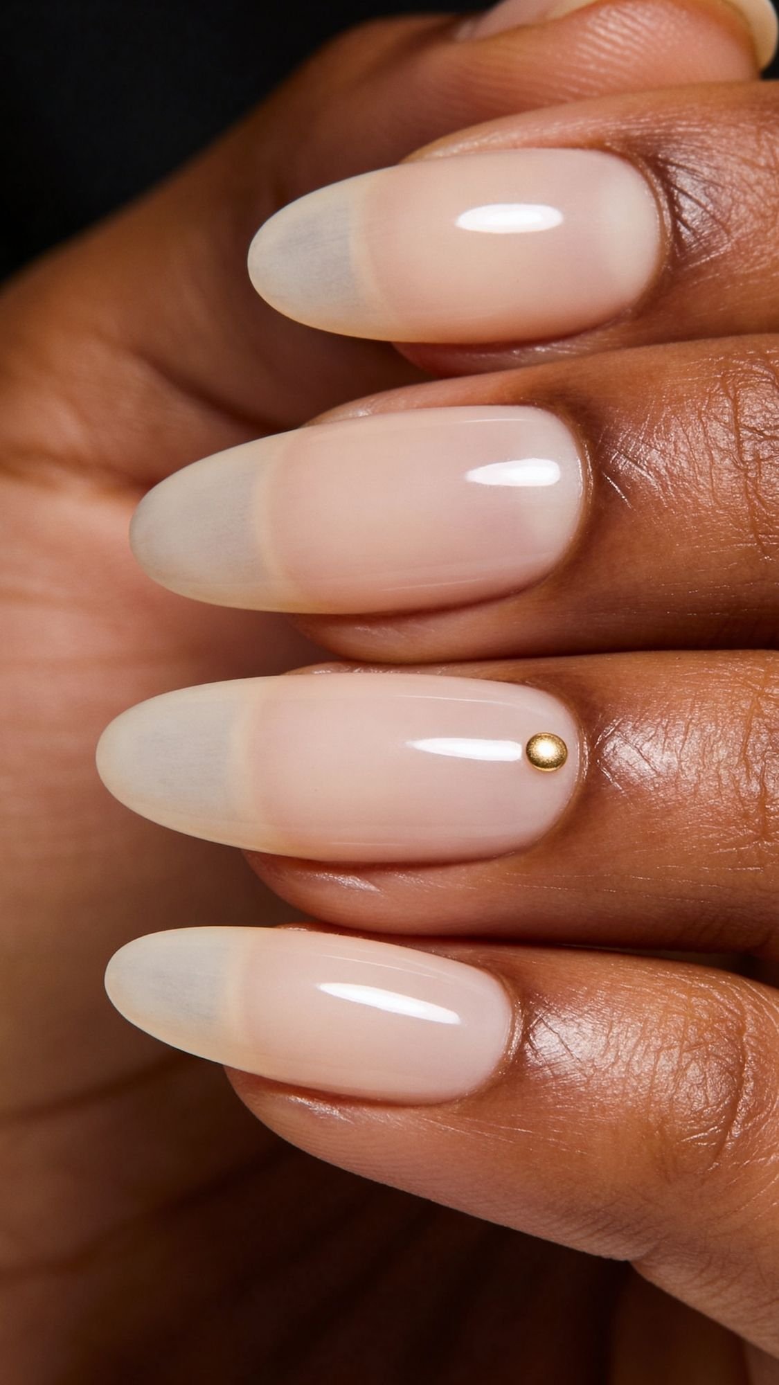 A close-up of a hand with long, almond-shaped, nude-colored nails—a polished everyday look. One nail features a small gold stud near the cuticle, perfect for chic nail ideas. The glossy finish adds to these clean girl nails.