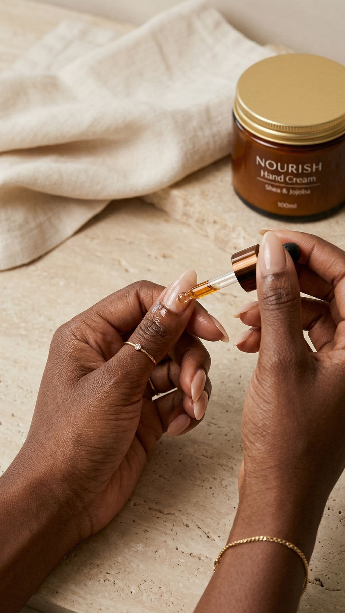 A person applies oil to their fingernail using a dropper—perfect for those seeking nourishing care for almond nails. In the background, a jar labeled “NOURISH Hand Cream” with shea and jojoba sits on a beige surface with fabric.