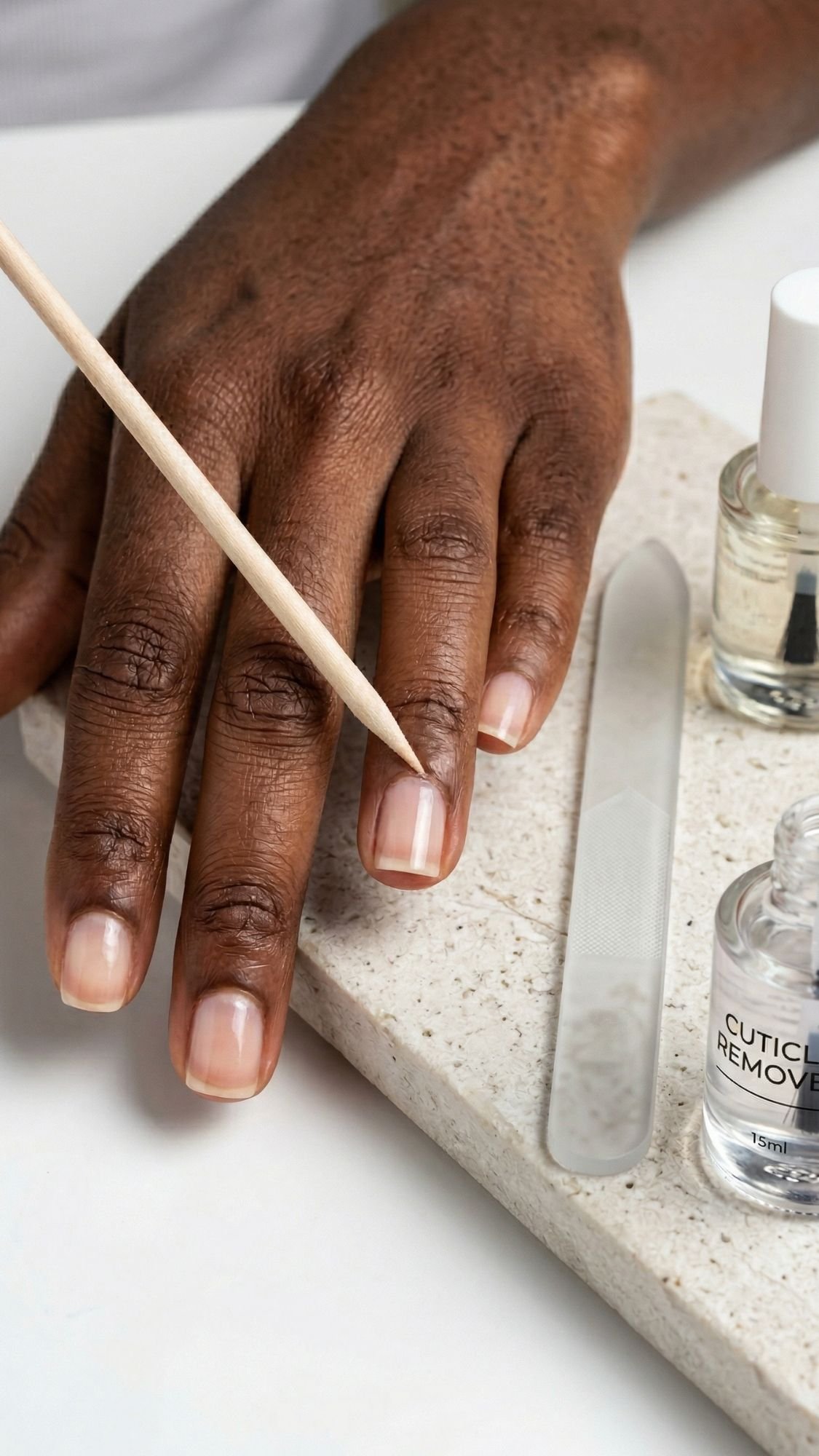 A hand is having its cuticles pushed back with a wooden stick. Nearby, a nail file, cuticle remover, and clear polish on a stone tray help create polished nails perfect for an everyday look or clean girl nails style.