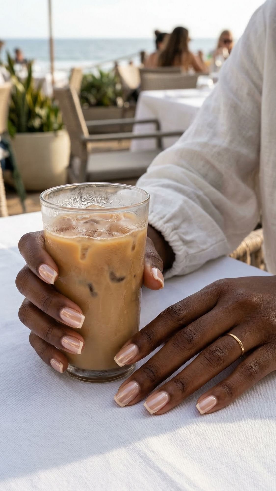 A person with neatly manicured summer nails holds a glass of iced coffee at an outdoor beachside café. Other people sit at tables in the background, with plants and the ocean visible.