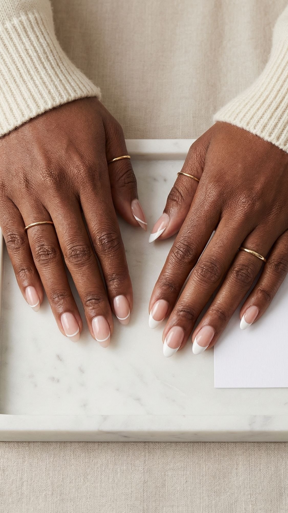 Two hands with almond-shaped French manicure rest on a marble tray, showing off flattering nails. The person wears thin gold rings on several fingers and a cream-colored sweater. A white card is partially visible on the tray.
