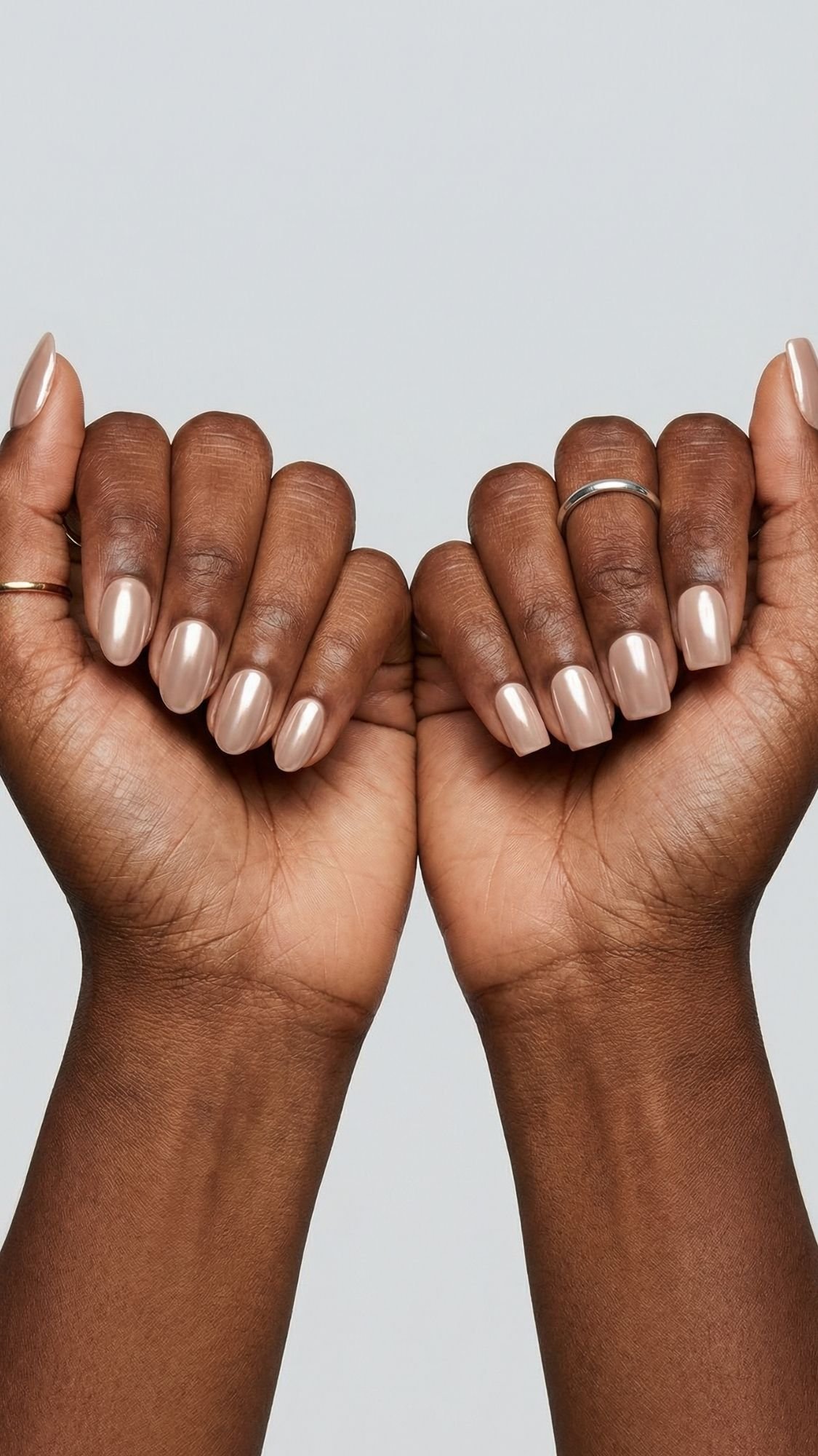 Two hands with dark skin are shown with fists facing forward. The glossy, light beige polish gives a subtle light reflecting nails effect. Both hands have thin, simple rings on the fingers against a plain light background.