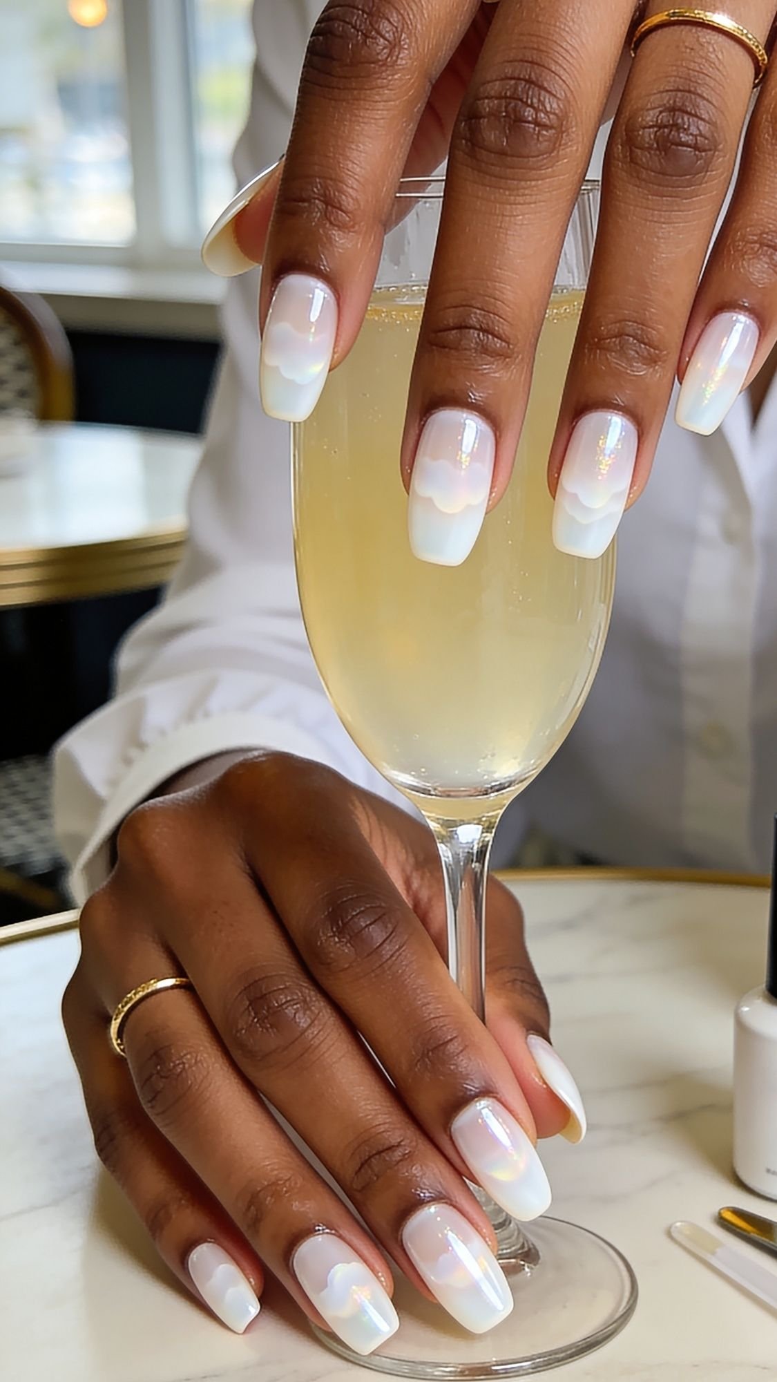 A person with medium brown skin and neatly manicured, iridescent white French tips—perfect summer nails—holds a champagne glass on a marble table. They wear gold rings and a white shirt, with a nail polish bottle visible nearby.