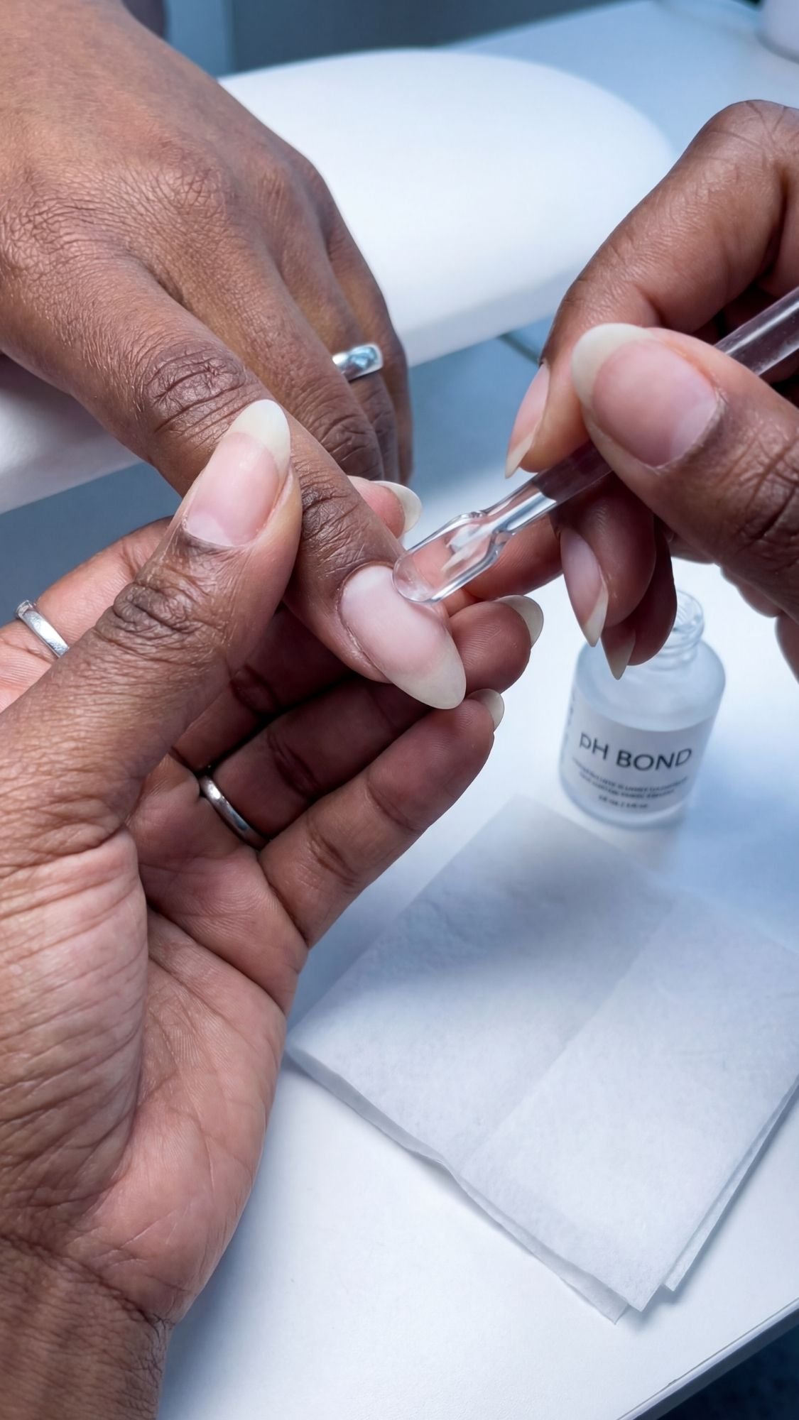 A close-up of hands during a manicure. One person applies pH Bond nail prep liquid to another’s natural nails, prepping for stunning Easter holiday nails, with a white towel and a bottle of pH Bond in the background.