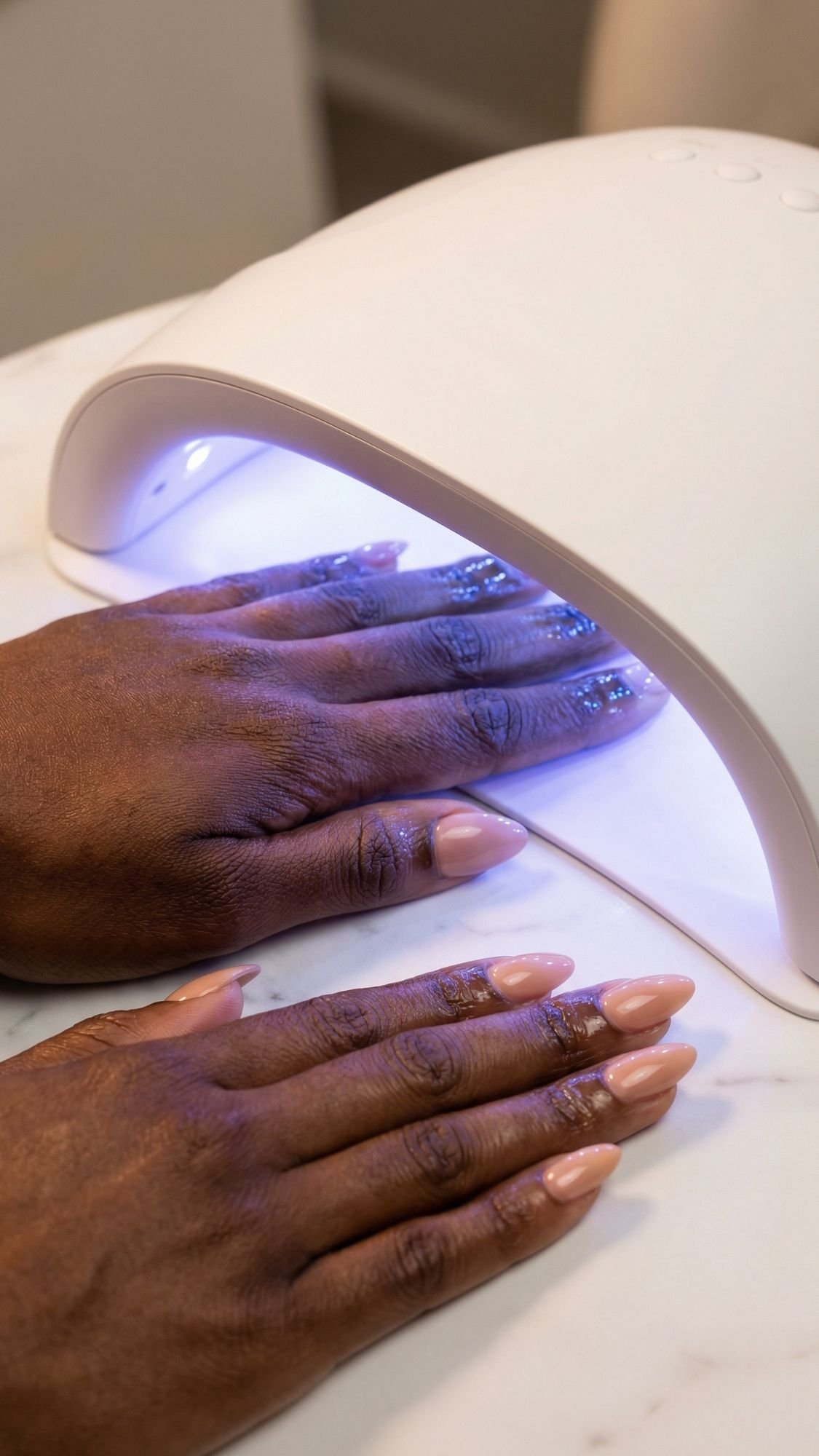 Two hands with freshly manicured, nude-colored nails—perfect for work friendly nails—are placed under a UV nail lamp, which emits a soft blue light during the gel nail curing process.
