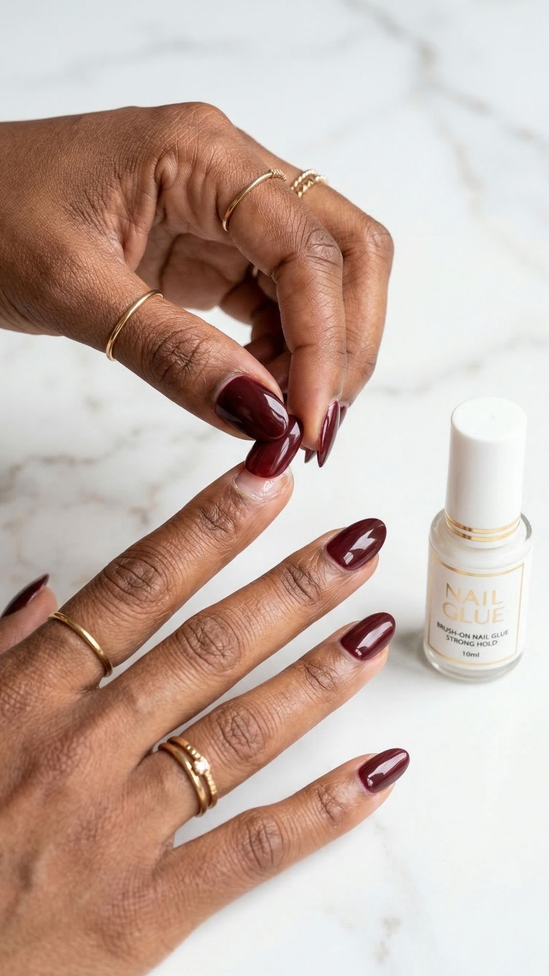 A person with dark skin and gold rings applies a burgundy press-on nail to a finger, creating elegant nails. A bottle of nail glue sits nearby on a white marble surface.