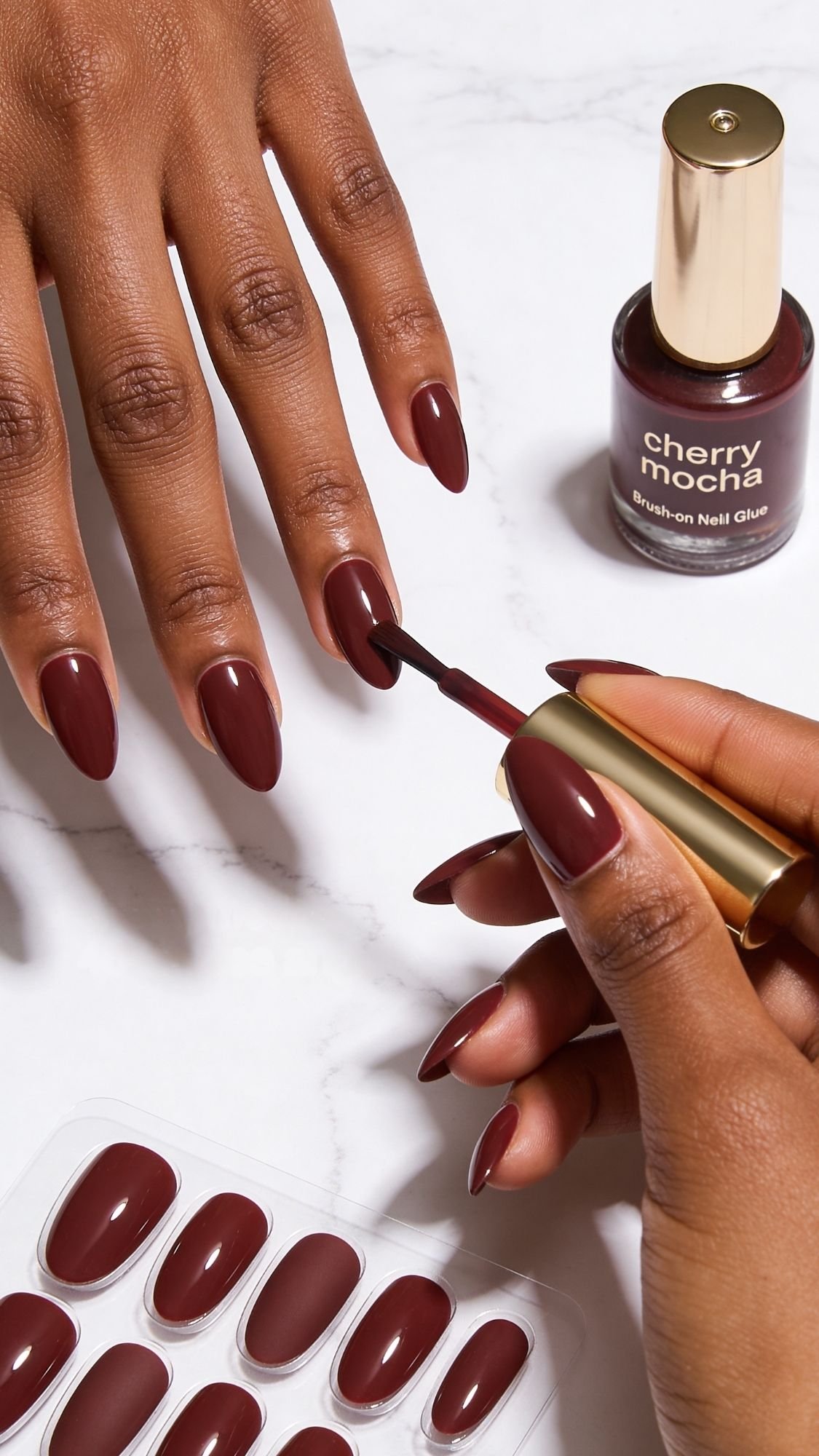 A person applies deep burgundy cherry mocha nail polish to their short nails. A bottle of polish and a set of matching press-on nails for elegant nail designs rest nearby on a white surface.
