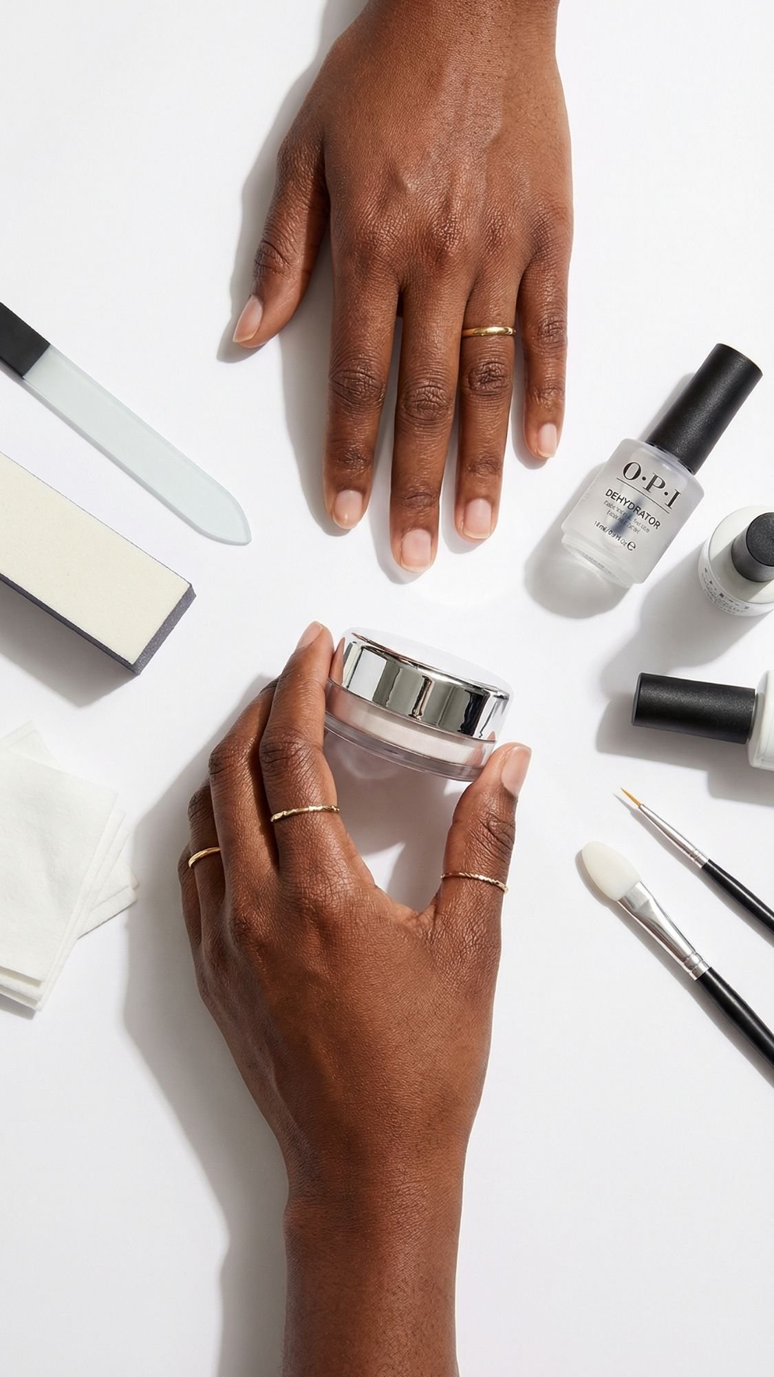 A person’s hands with simple gold rings are surrounded by manicure tools, a nail file, polish bottles—including chrome nails—brushes, buffers, and a small jar, preparing for a nail care routine on a light white surface.