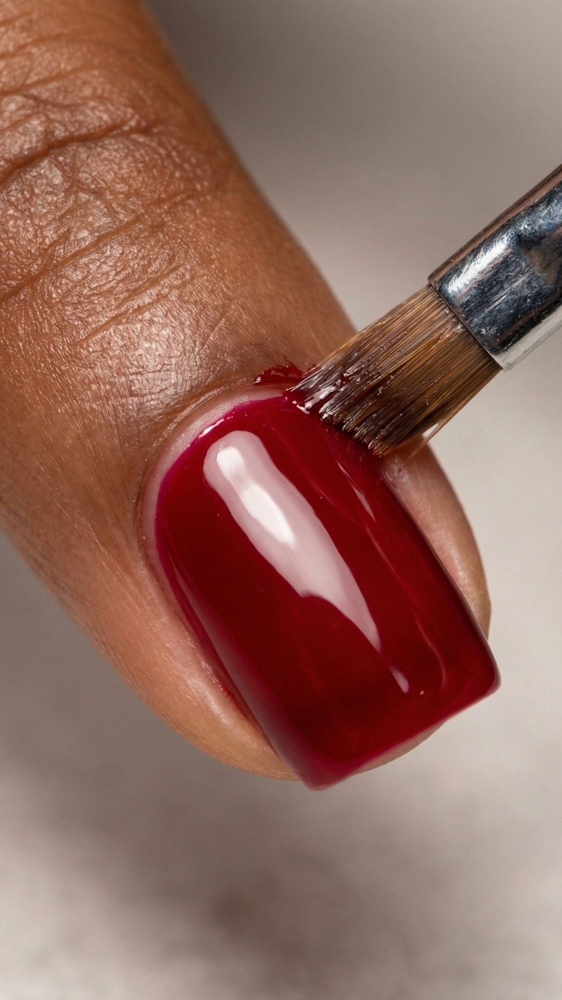 A close-up of a hand painting a fingernail with glossy red nail polish using a small brush. The nail polish is being carefully applied to the brown-skinned person's nail, perfect for chic summer nails.