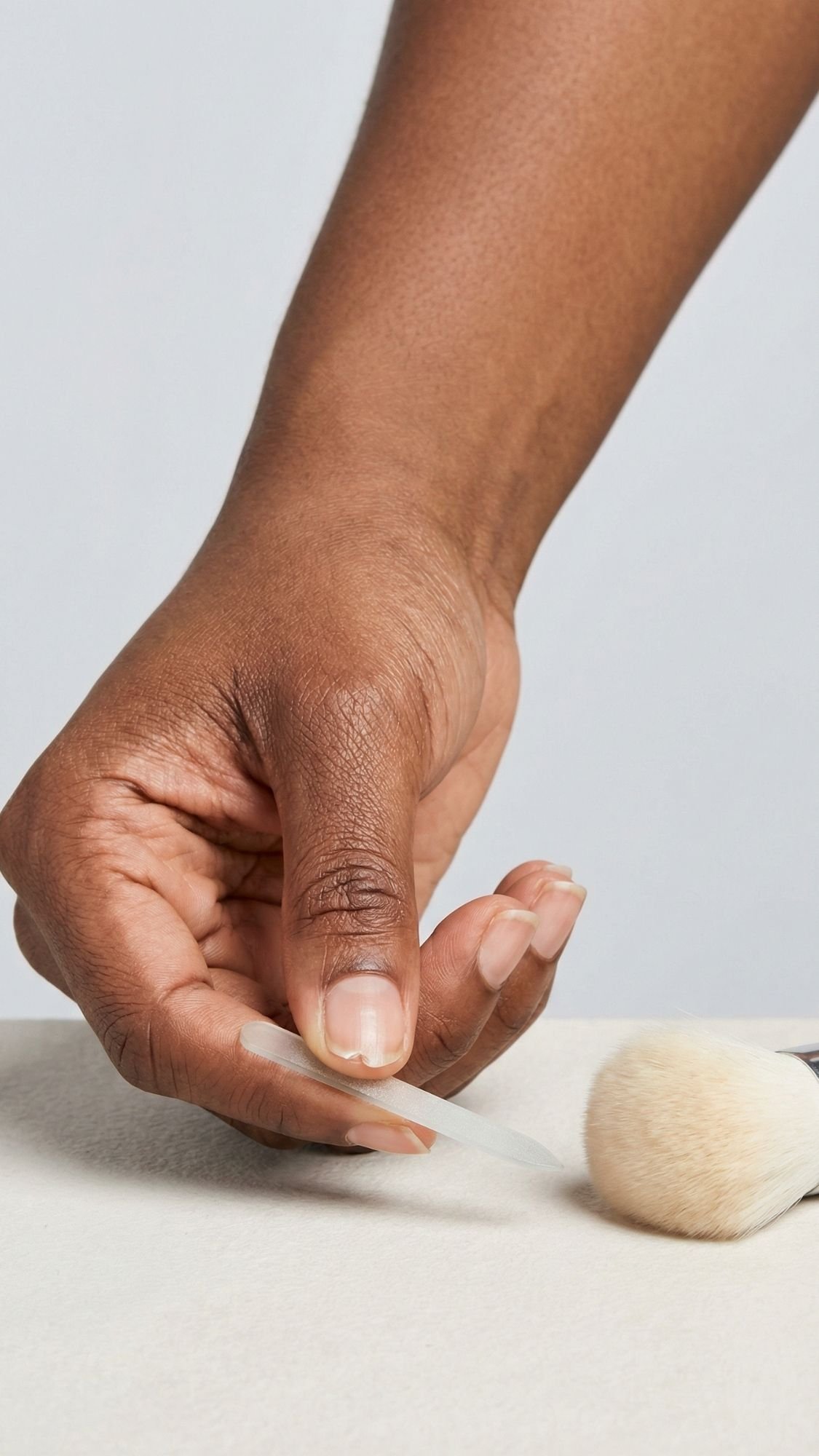 A close-up of a person’s hand with natural nails holding a nail file, next to a makeup brush resting on a light surface—perfect prep for creating stunning Chrome Nail Designs.