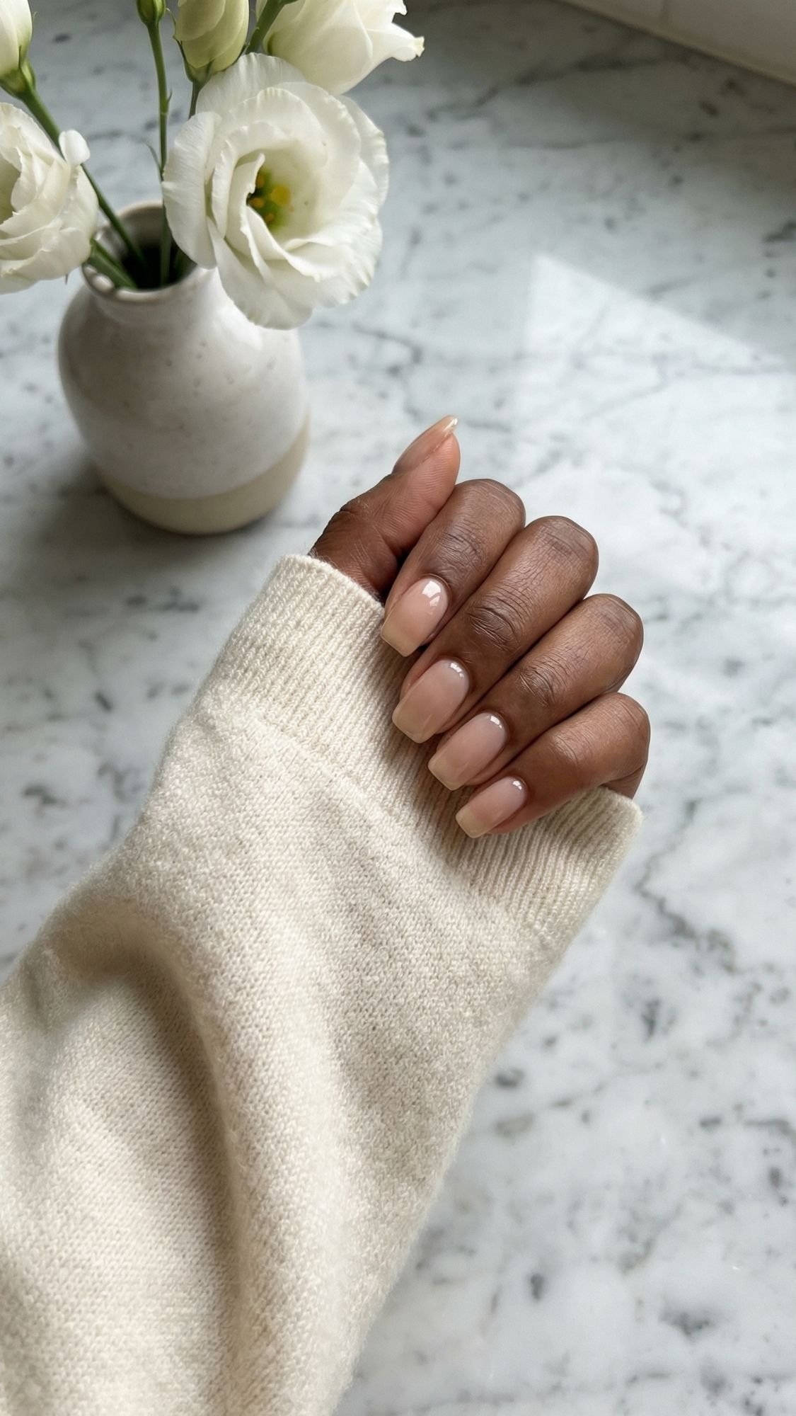 A person with medium brown skin shows off polished nails in a natural nude shade, wearing a cream sweater. A white ceramic vase with white flowers sits on a marble surface in the background, completing this effortless everyday look.