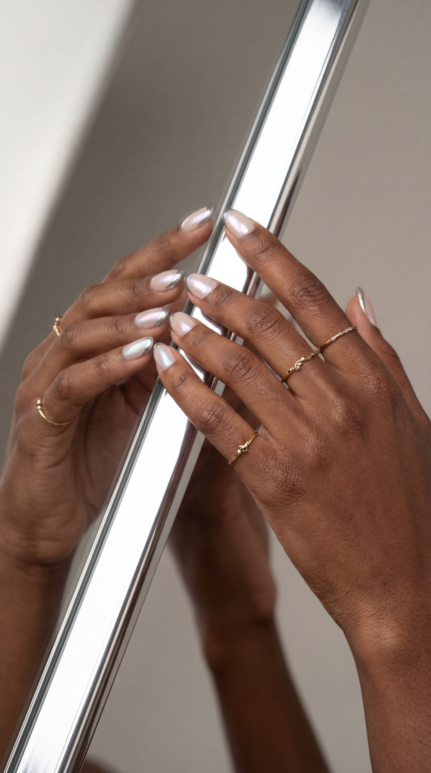 A close-up of two hands with light pink polished nails and several delicate gold rings, gently touching a reflective metallic surface, evoking chic Chrome Nail Designs as the mirror shows the reflection of the hands and rings.