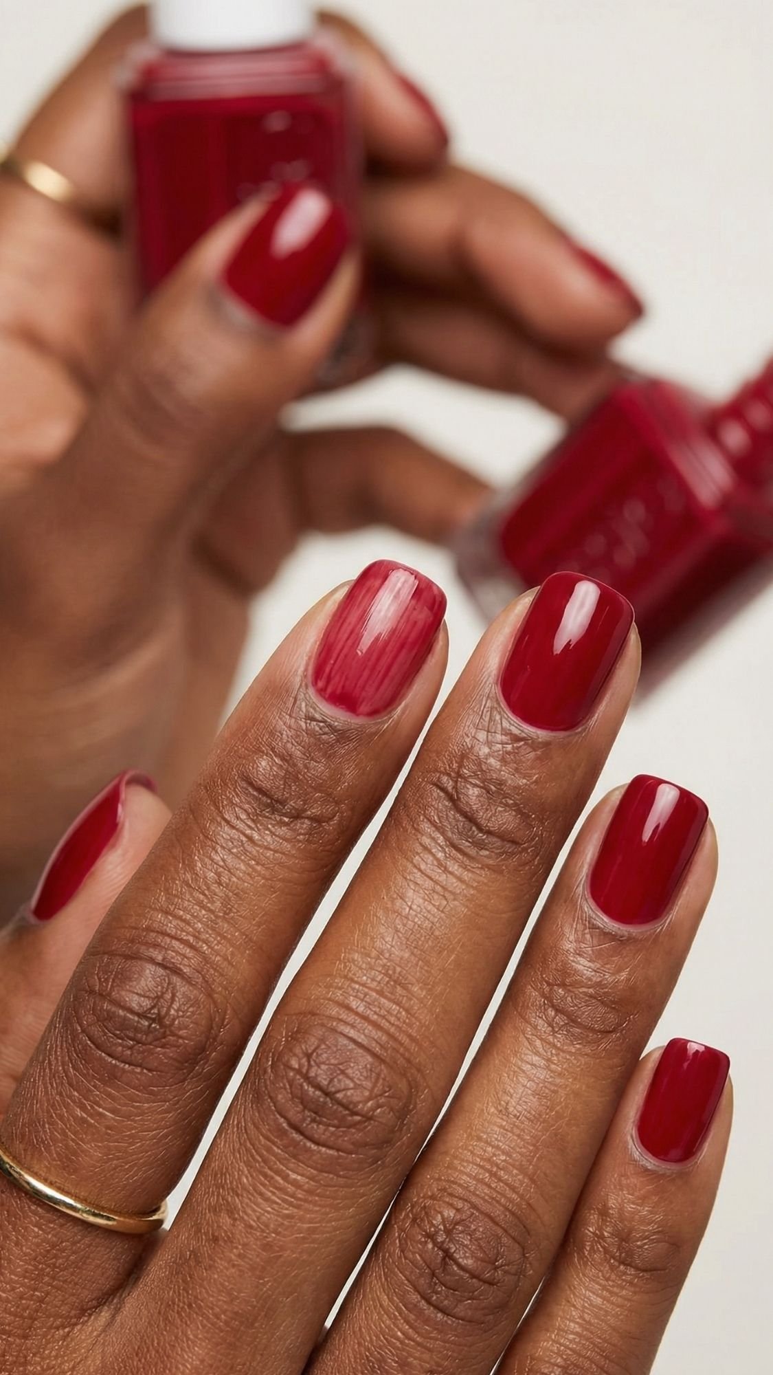 A close-up of a hand with neatly manicured, glossy red nails being painted—a perfect pick for seasonal nails. Another hand holds the bottle in the background, while thin gold rings add an elegant touch.