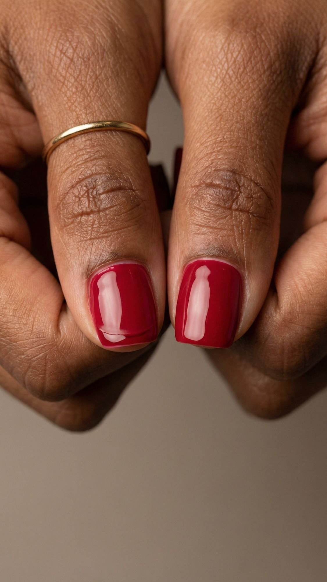 Close-up of two thumbs with short, neatly manicured red nails—an elegant nail idea for every season. One thumb is adorned with a simple gold ring, set against a neutral, softly blurred background.