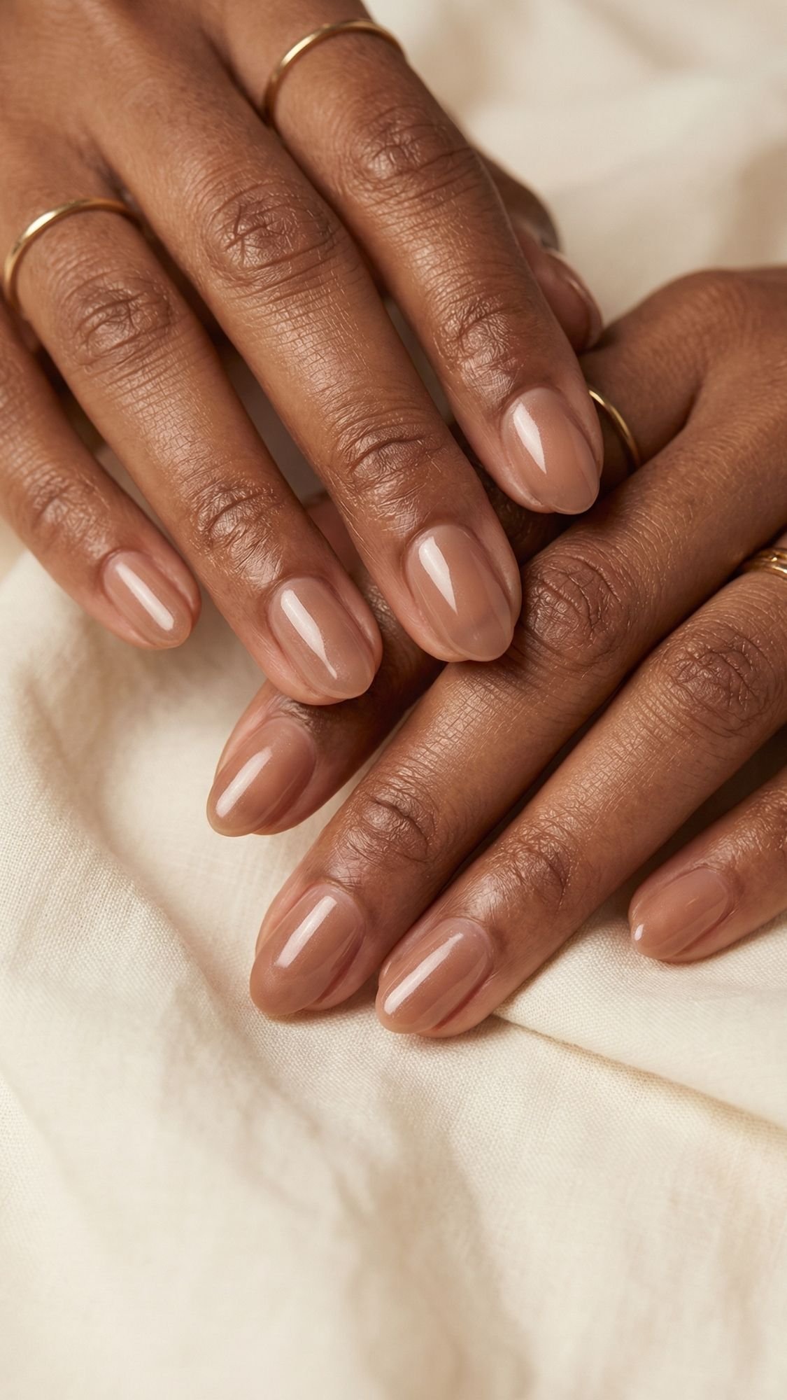 Close-up of hands with smooth, medium-brown skin resting on cream fabric. Nails are almond-shaped and painted with glossy nude polish—perfect inspiration for summer nails. The fingers are adorned with slim gold rings.
