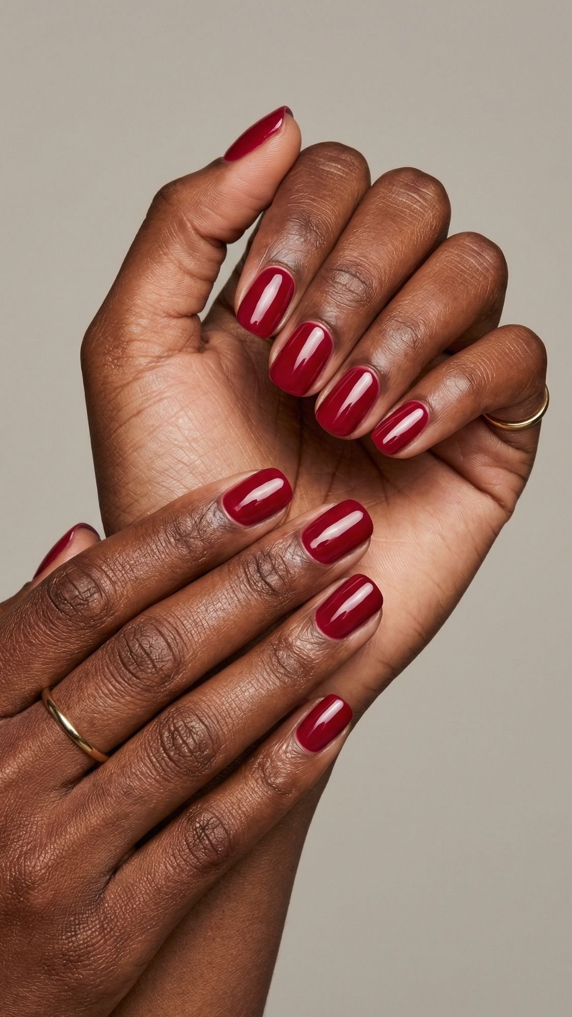 Two hands with dark skin, wearing thin gold rings, show off neatly manicured red nails painted in glossy deep polish—a chic option for those seeking fresh nail ideas against a neutral background.