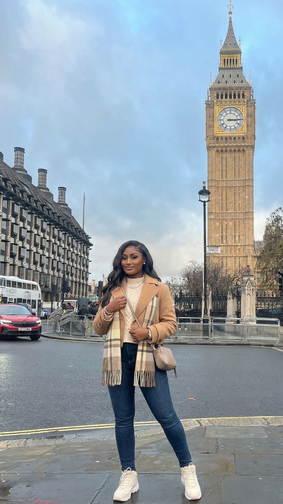 A woman in a tan coat, scarf, jeans, and white sneakers stands on a street near Big Ben—an iconic sight for anyone planning their first trip to London. The clock tower and historic buildings create the perfect London travel backdrop under a cloudy sky.